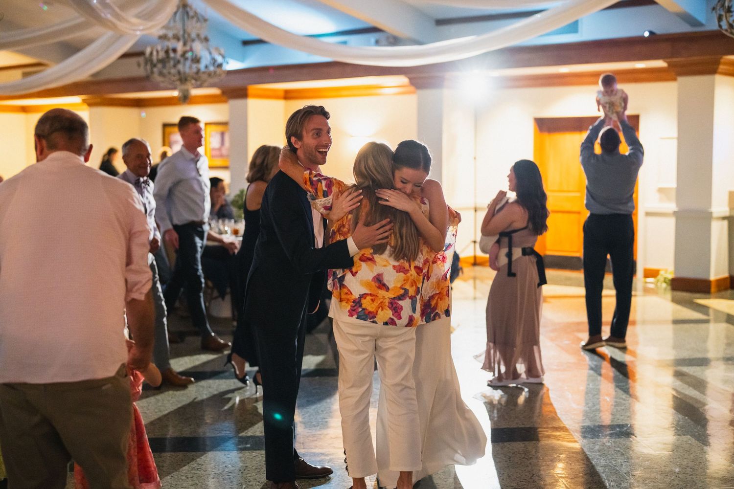 Wedding guests embrace and celebrate on the dance floor during a lively indoor reception party.