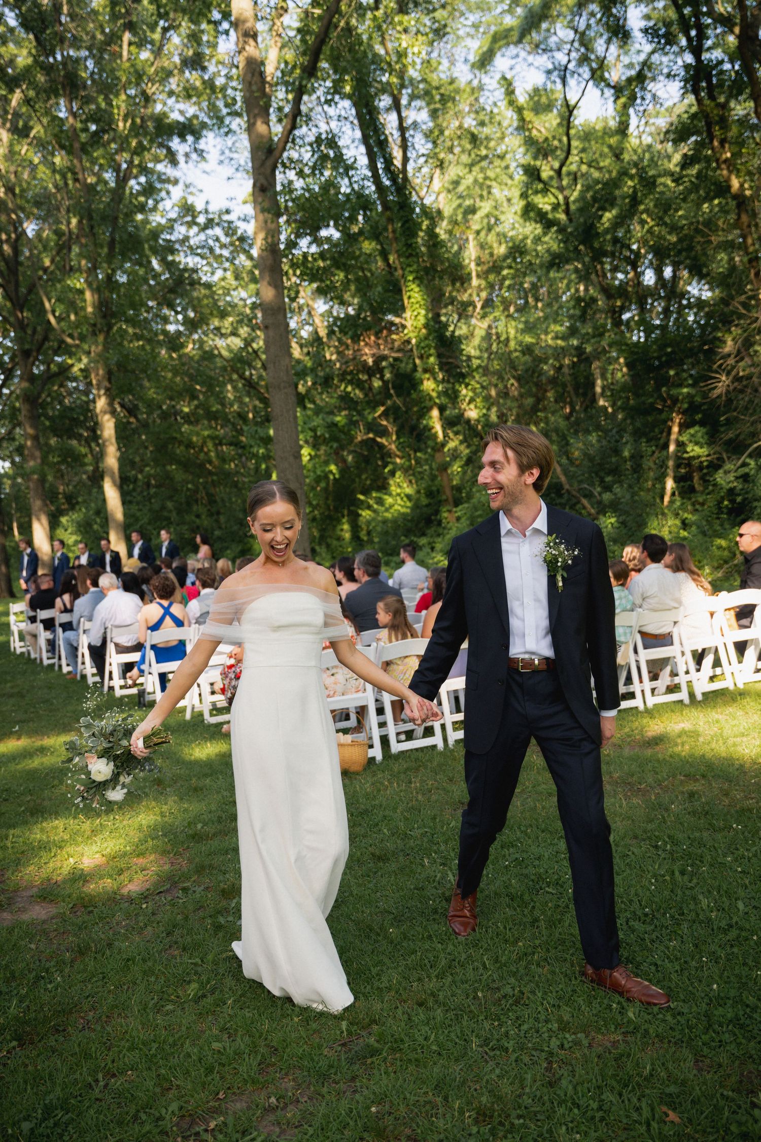 Newlyweds walk hand-in-hand down the aisle at an outdoor woodland wedding ceremony.
