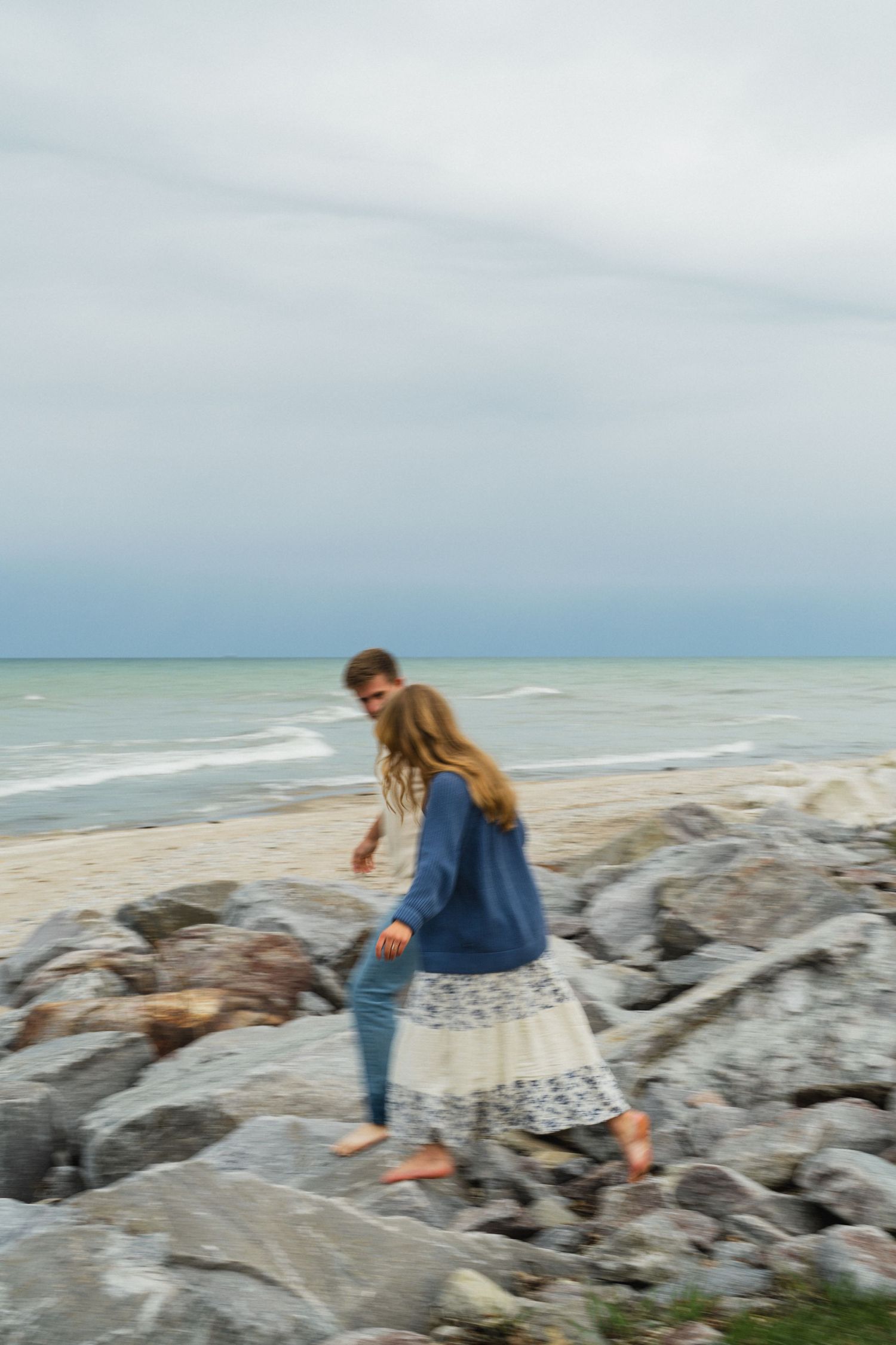 Couple walking barefoot on rocky beach shoreline, woman in lace skirt and denim jacket, motion blur effect.