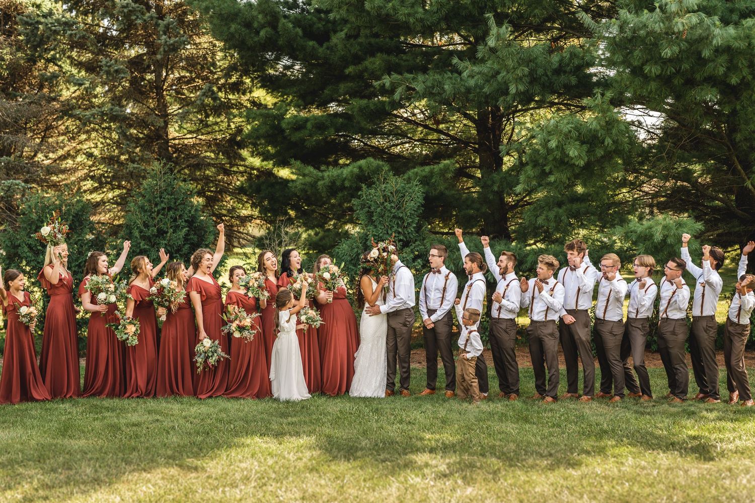 Wedding party cheers and celebrates on grassy lawn with pine trees in background.