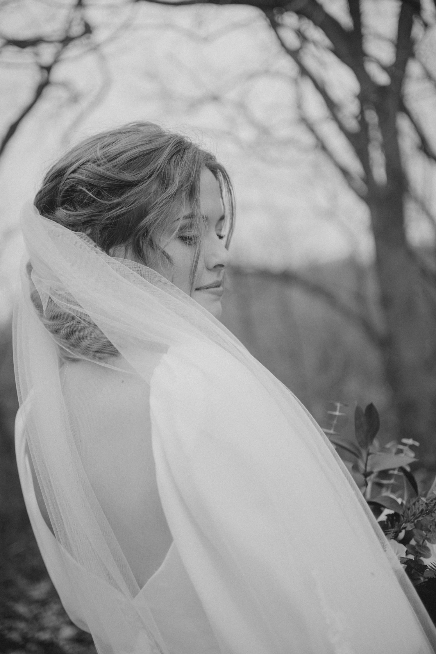 Black and white portrait of a bride with flowing veil looking down in a natural outdoor setting.