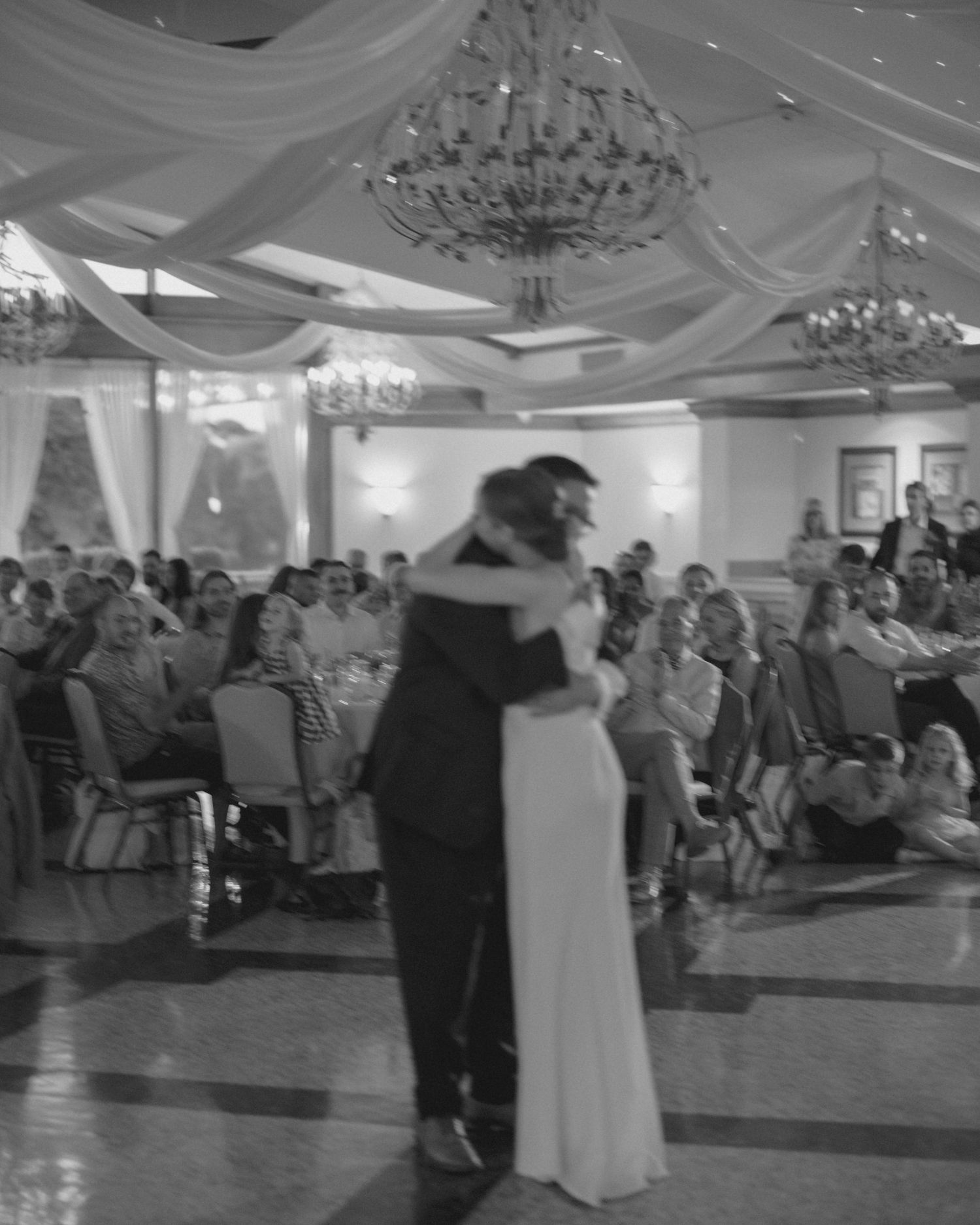 Bride and groom share first dance in black and white photo before guests in grand ballroom.