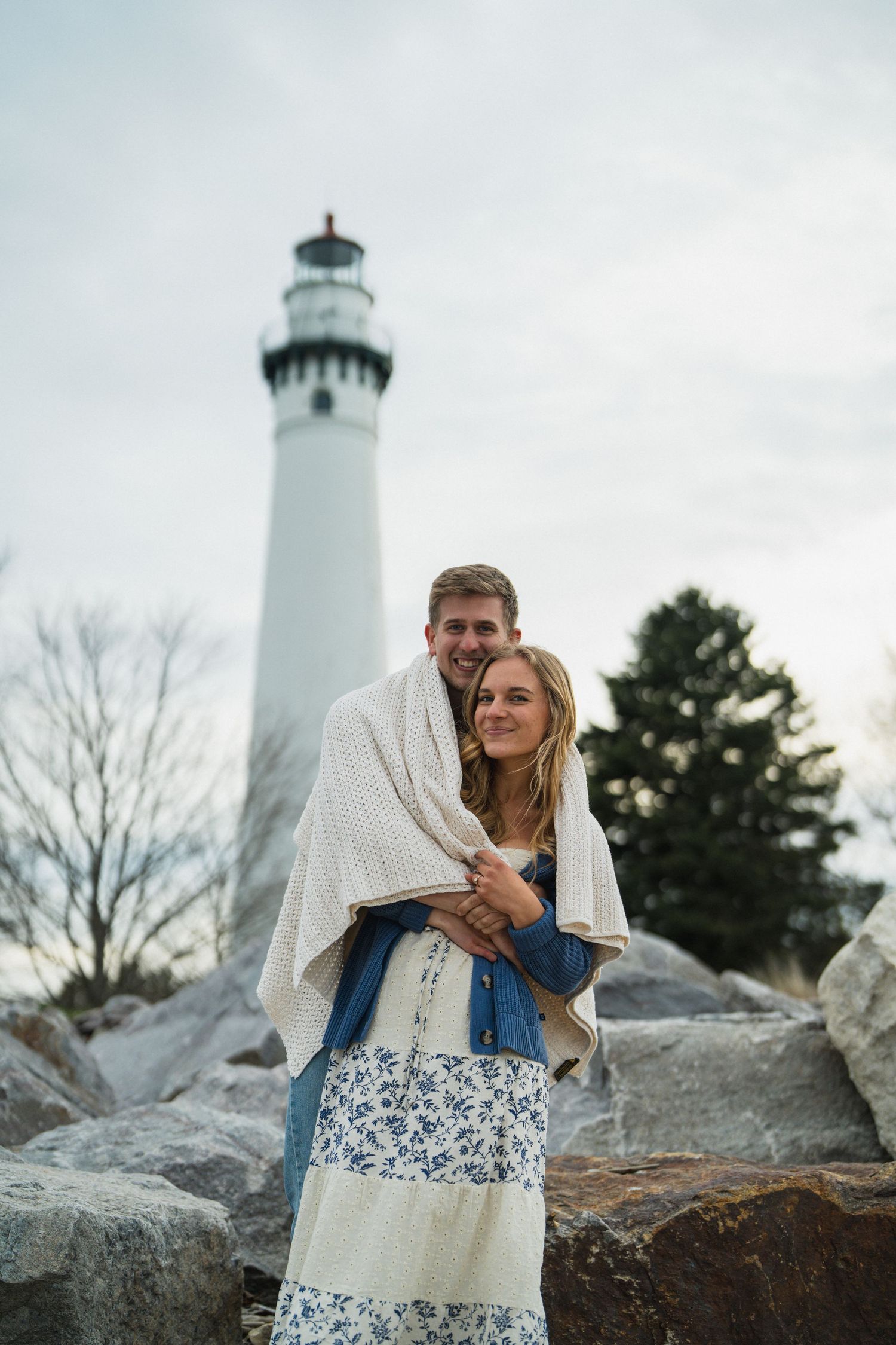 Couple wrapped in blanket standing on rocks before a white lighthouse on a cloudy day.