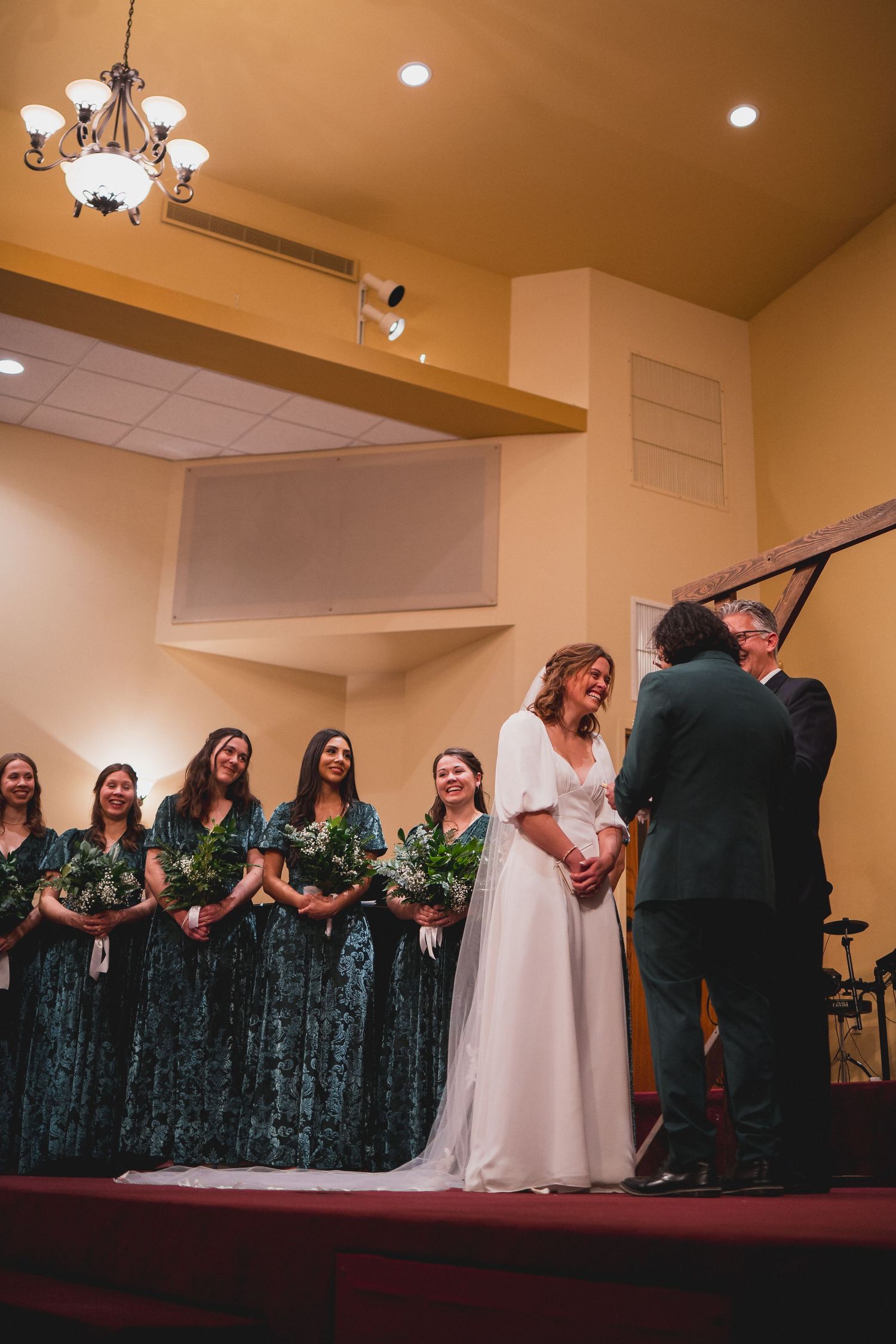 Wedding party arranged on church stage with bridesmaids in dark dresses.