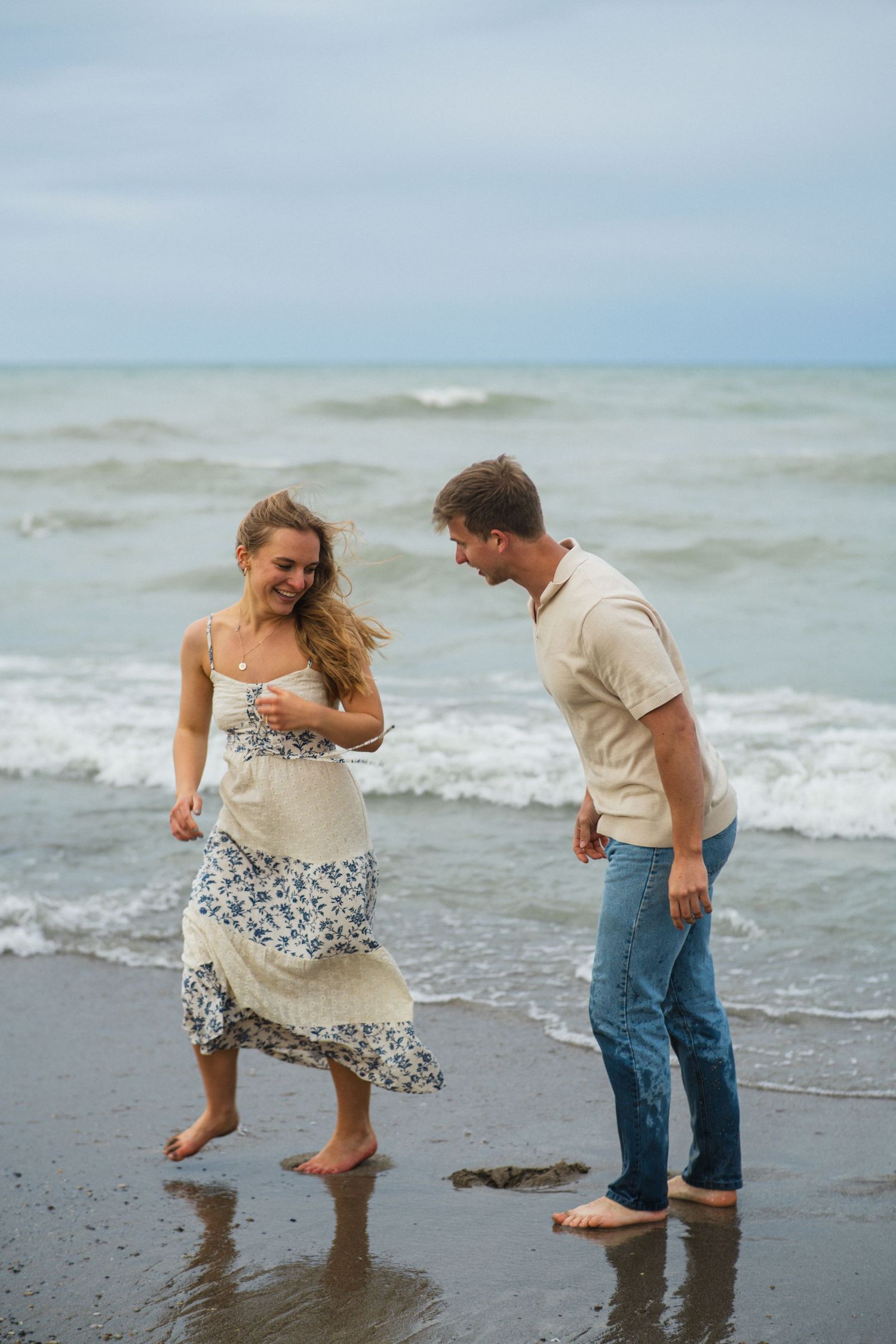 Happy couple laughing and dancing barefoot on a sandy beach with ocean waves behind them.