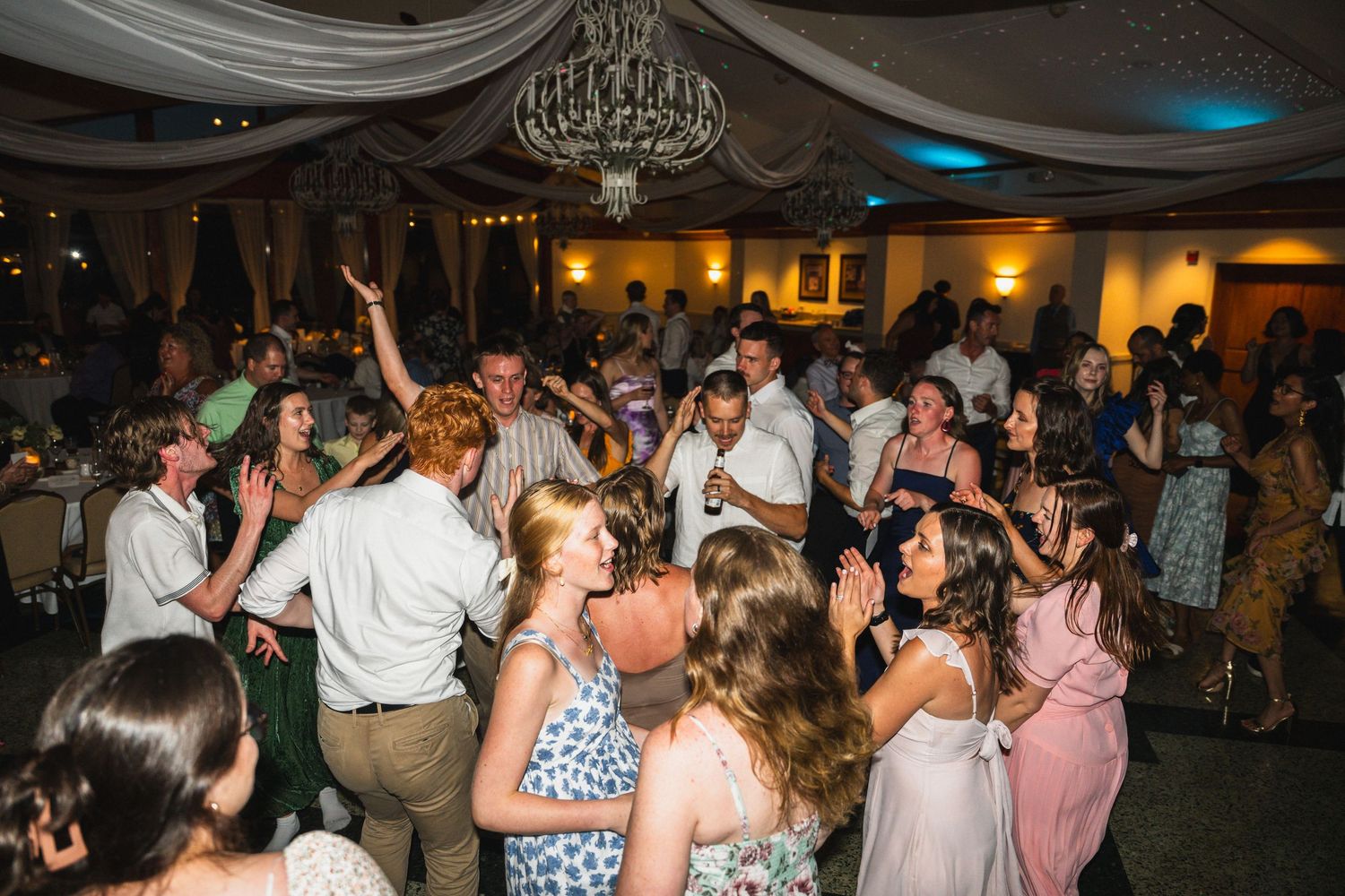 Energetic wedding reception dance floor packed with celebrating guests under chandelier.