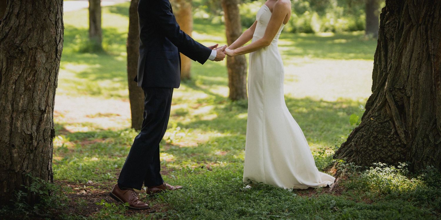 Bride and groom hold hands between two large trees in a sunlit park setting.