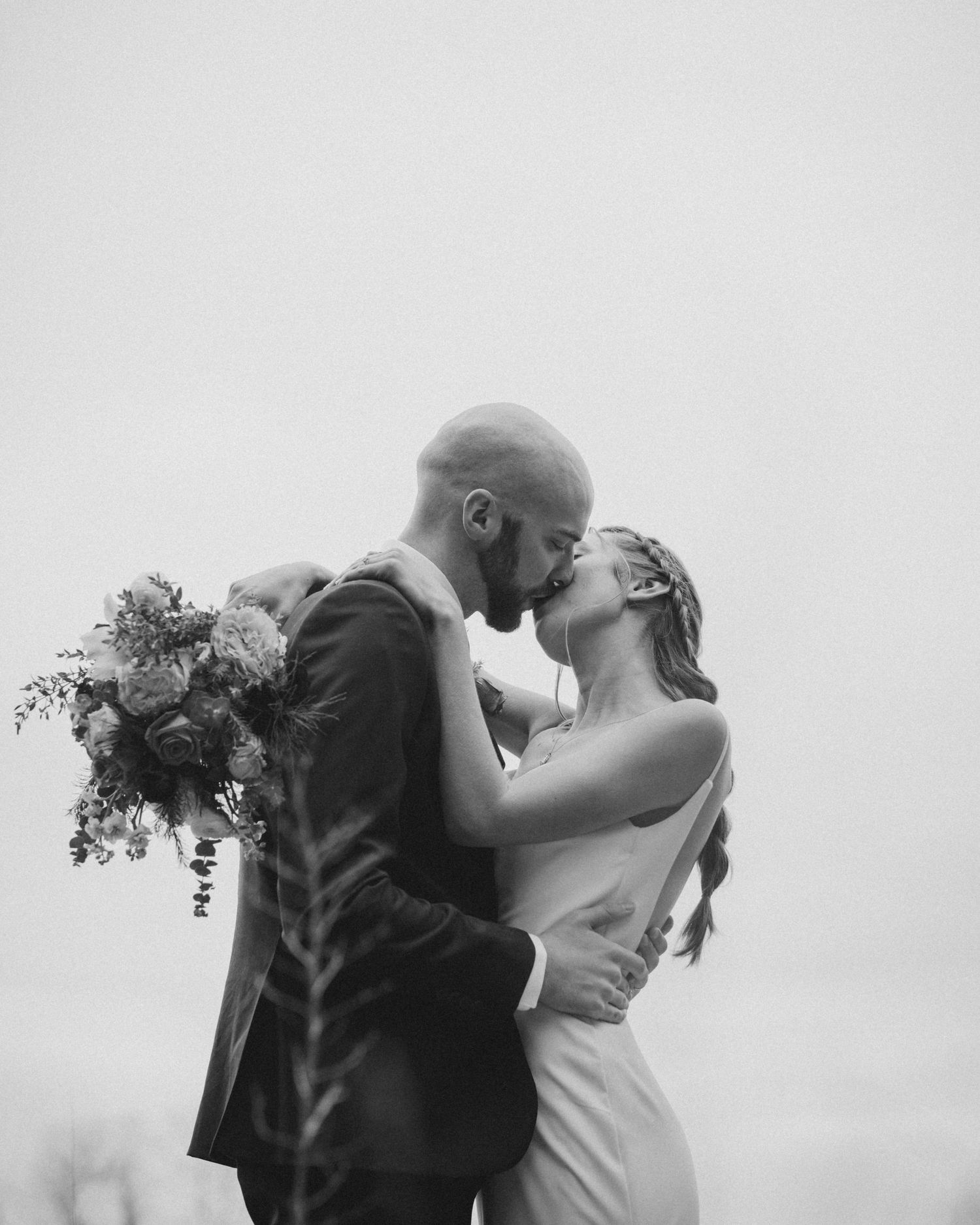 A tender moment between newlyweds sharing a kiss against an overcast sky.