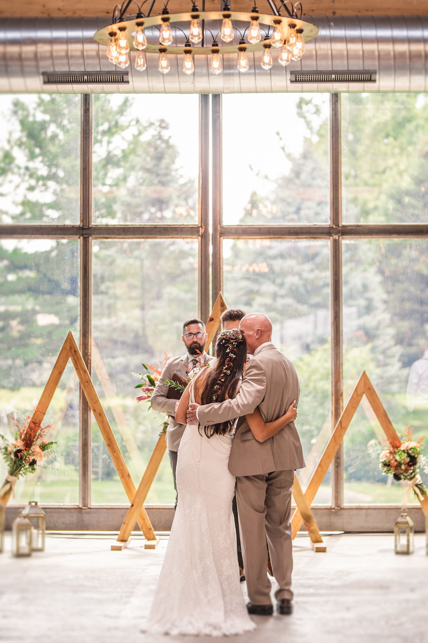 Wedding ceremony with wooden geometric arch and chandelier in an industrial-style venue with large windows.