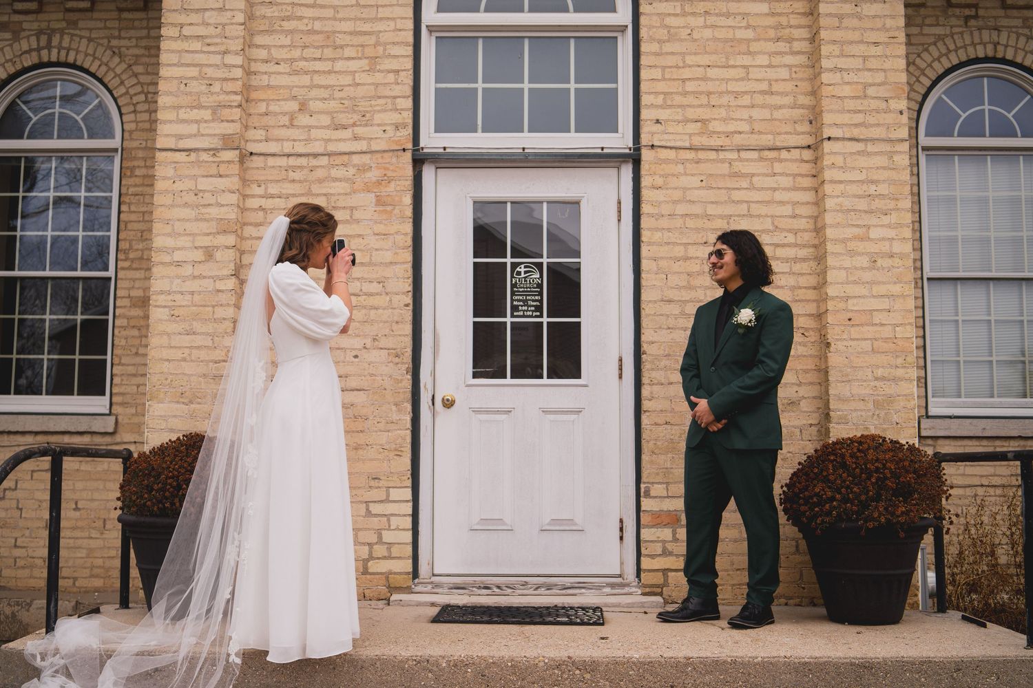 A wedding couple poses in front of a brick building with white door, bride in white dress and groom in green suit.