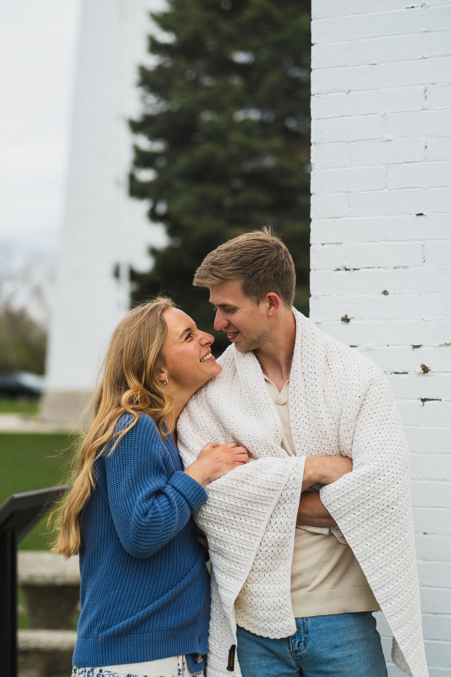 A smiling couple in cozy sweaters share a romantic moment leaning against a white brick wall outdoors.