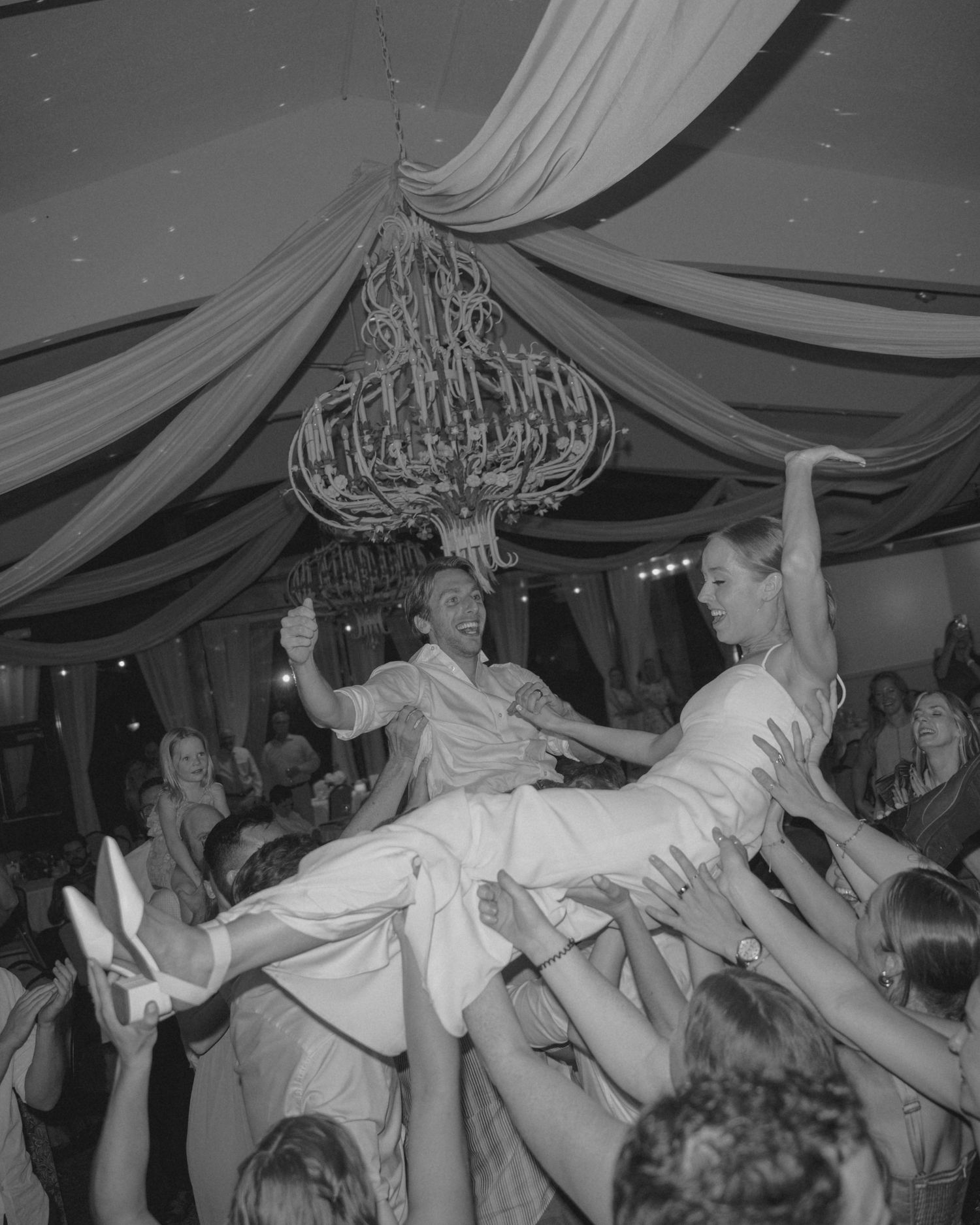 Black and white photo of bride and groom being lifted by wedding guests during lively reception dance.