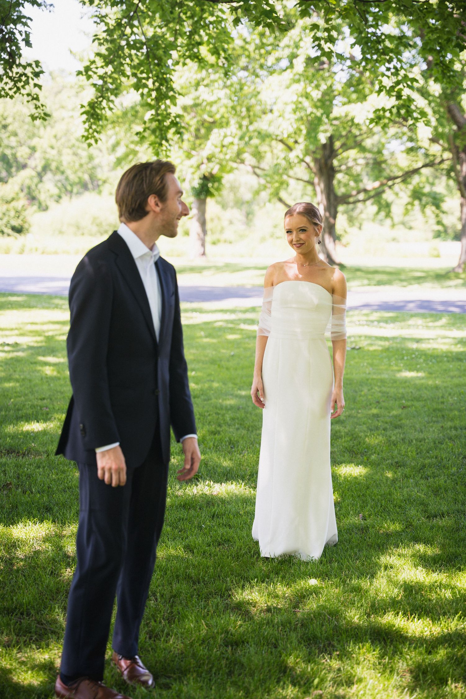 Groom in navy suit faces smiling bride in white off-shoulder gown during outdoor wedding first look in sunny park.
