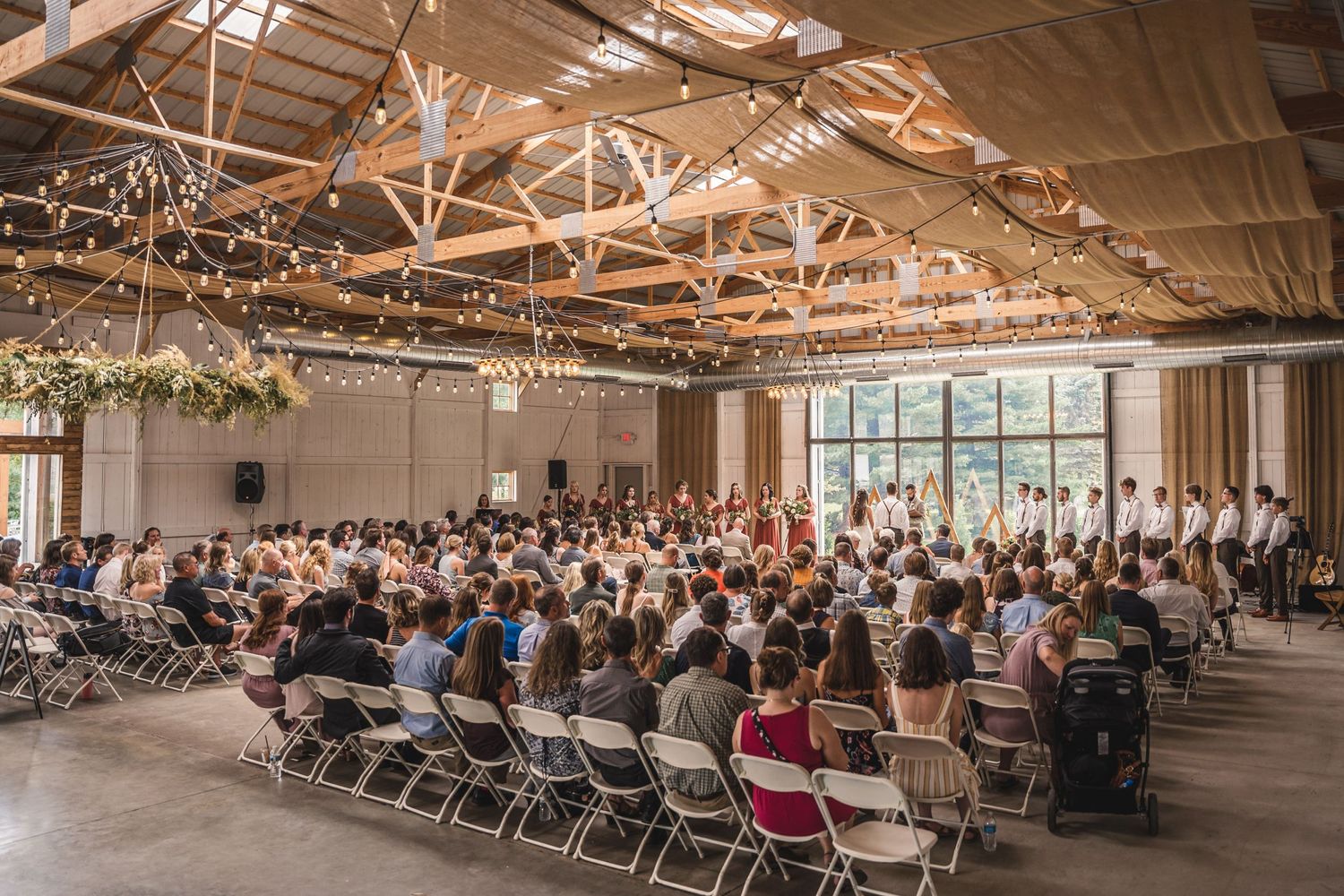 Large wedding ceremony venue filled with guests seated in rows of folding chairs under industrial ceiling.