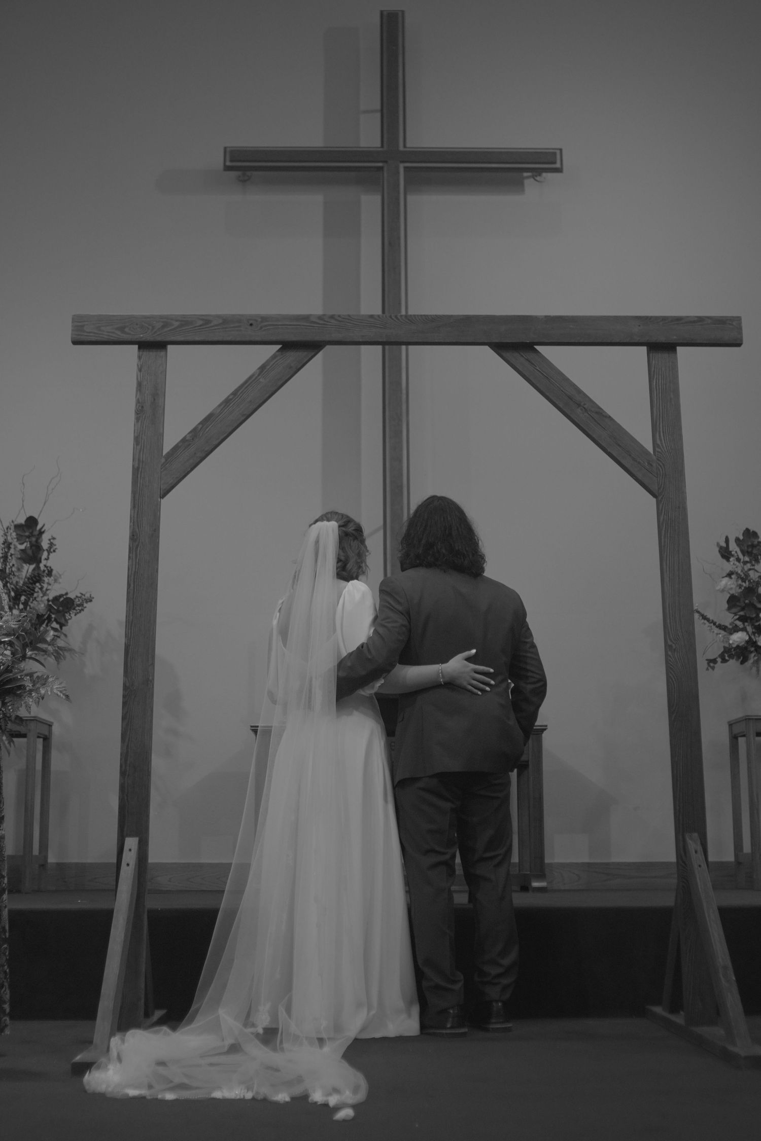A black and white photo of newlyweds embracing beneath a wooden cross altar.