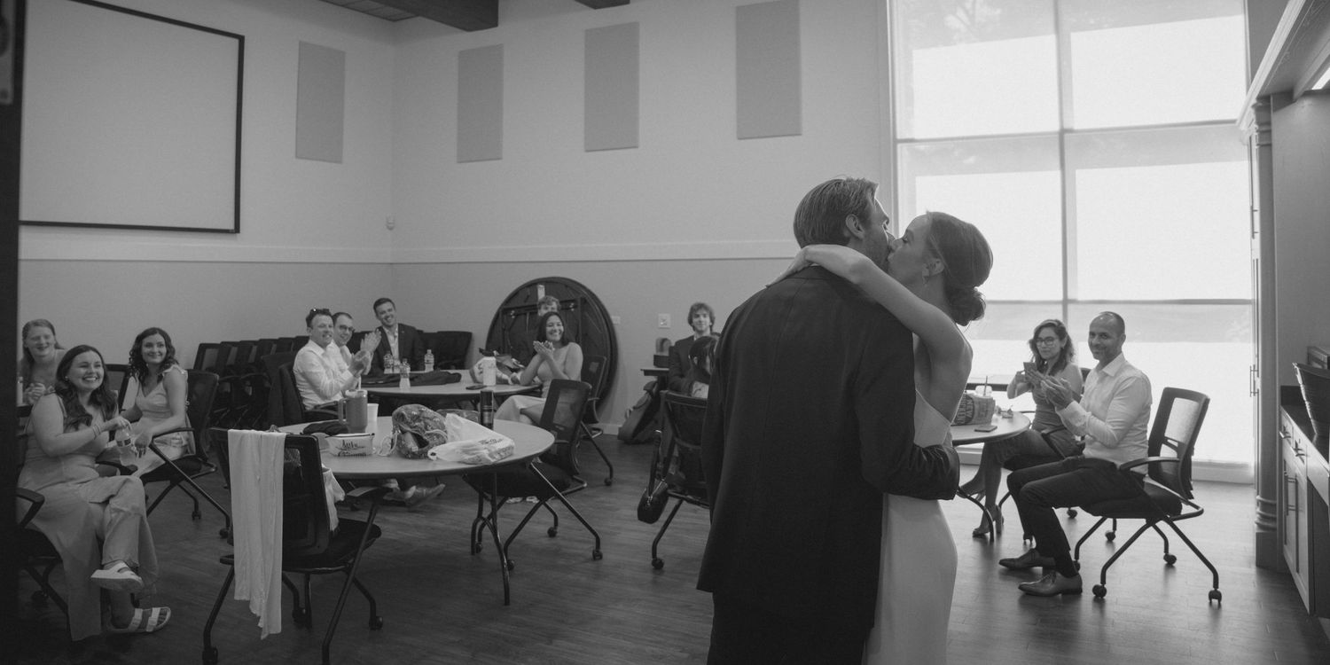Black and white photo of professionals in a modern meeting room, two people embracing while colleagues watch.