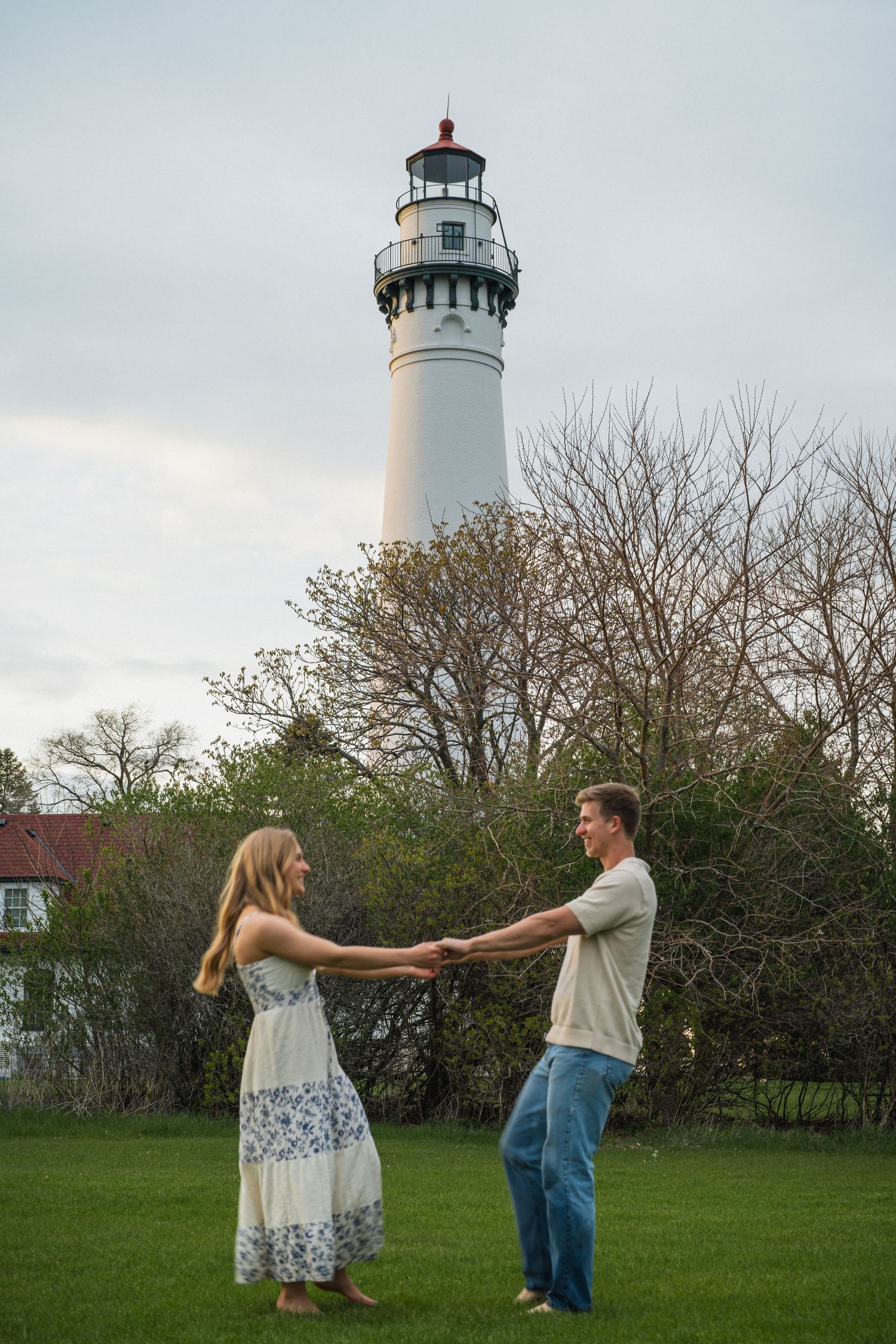 Couple holding hands facing each other on a lawn with a tall white lighthouse in the background.