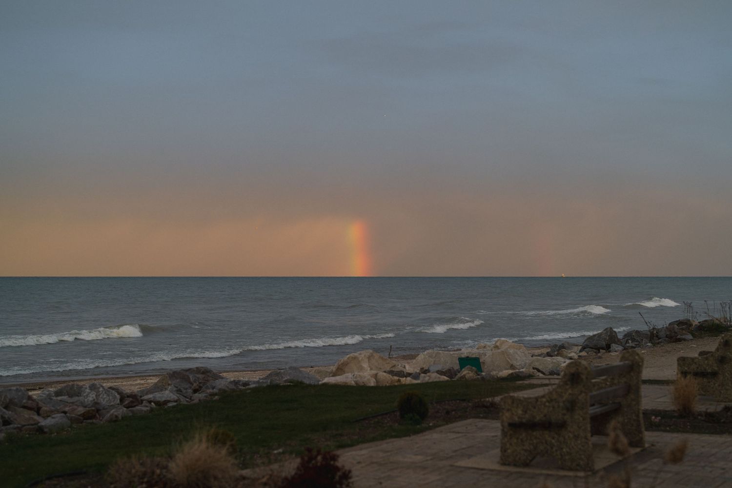 Scenic coastal sunset view with a faint rainbow over calm ocean waves and rocky shoreline.