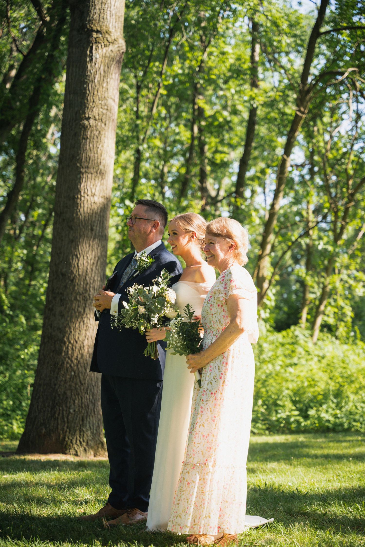 Bride stands between her parents in a sunny green park holding a white floral bouquet.