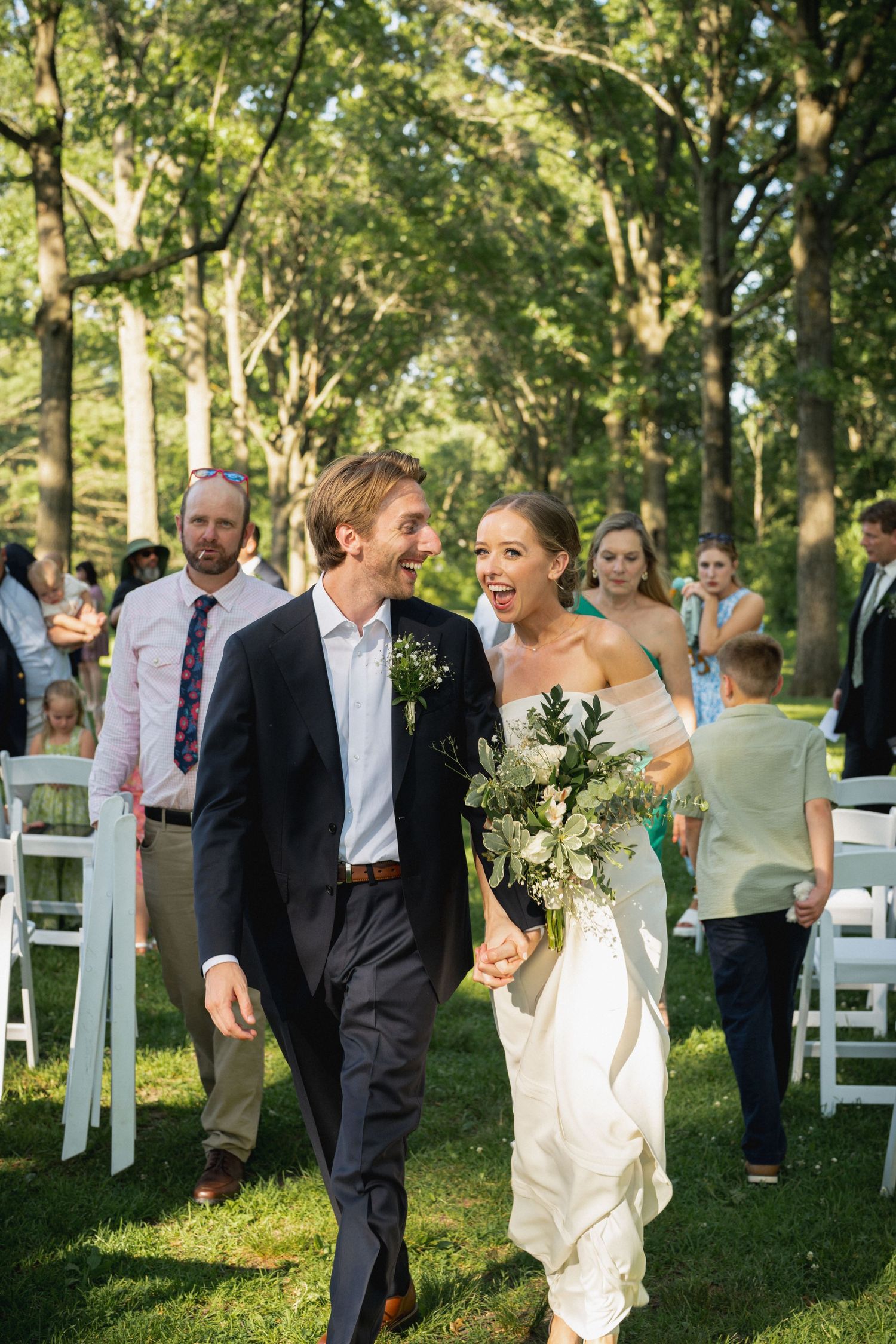 Joyful newlyweds walk down outdoor aisle holding hands, bride in white gown with bouquet, groom in navy suit, guests applaud.