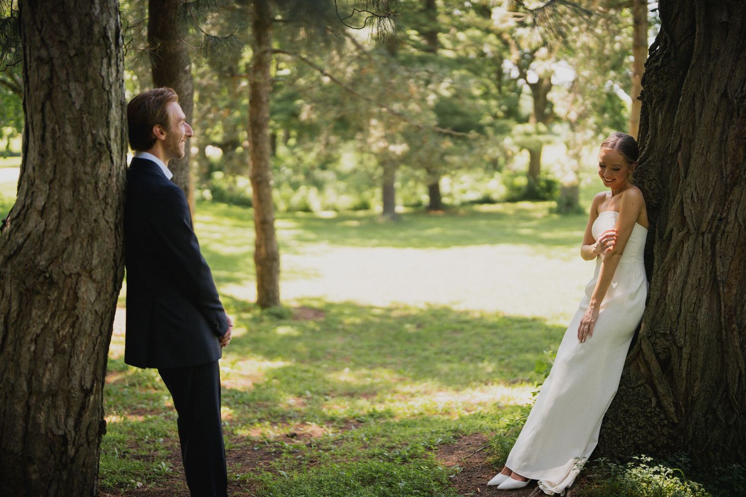 Wedding couple standing between tall trees in a sunlit forest setting.