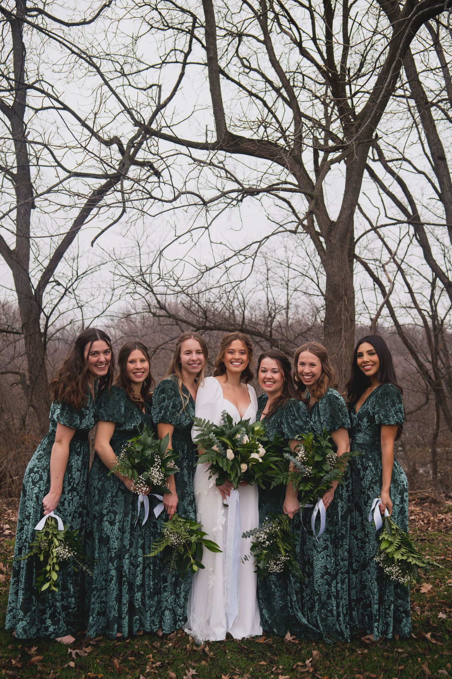 Bridal party in emerald velvet dresses poses beneath bare winter trees with white floral bouquets.