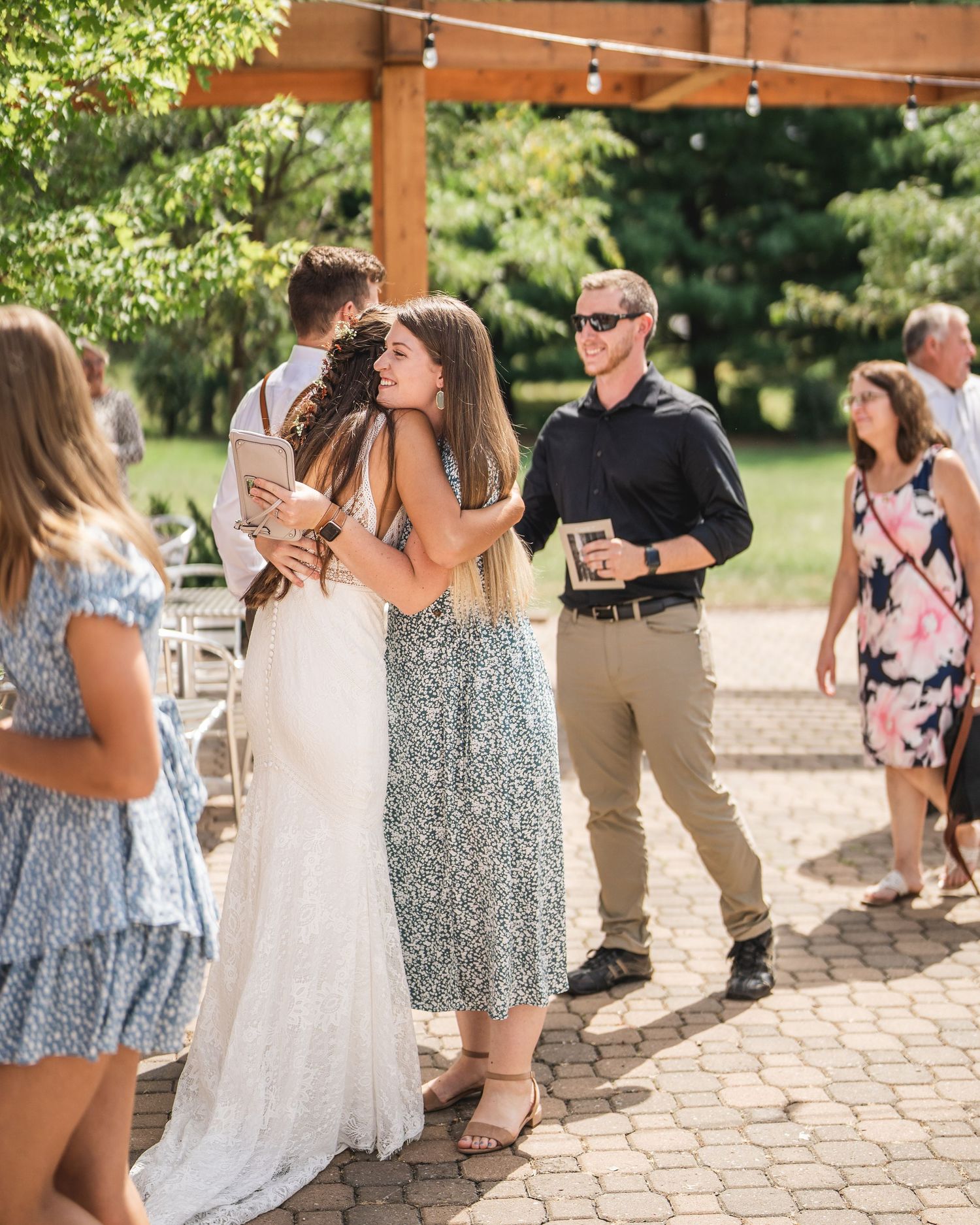 Wedding guests mingle on a brick patio under a wooden pergola during an outdoor reception celebration.