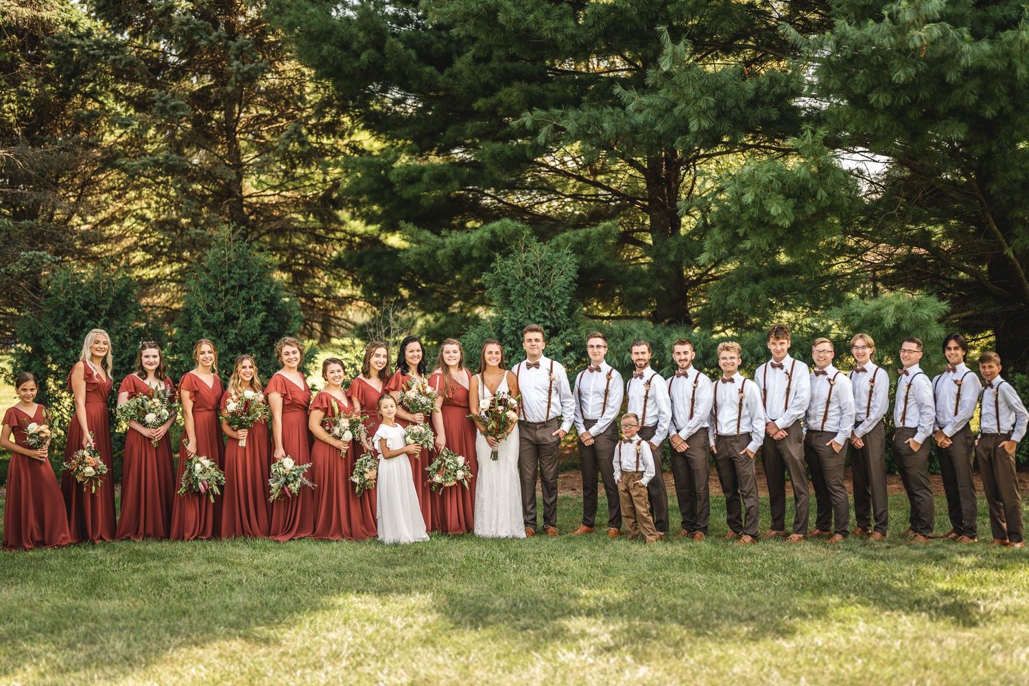 Wedding party in burgundy dresses and brown suspenders poses outdoors against evergreen trees.