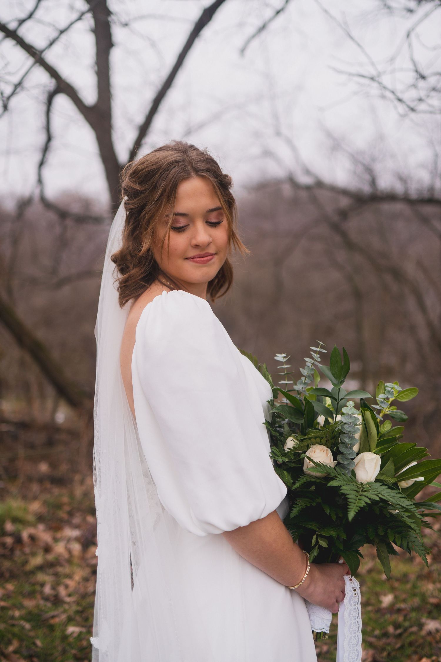 A bride in a white dress with puff sleeves holds a green and white bouquet in a wooded setting.
