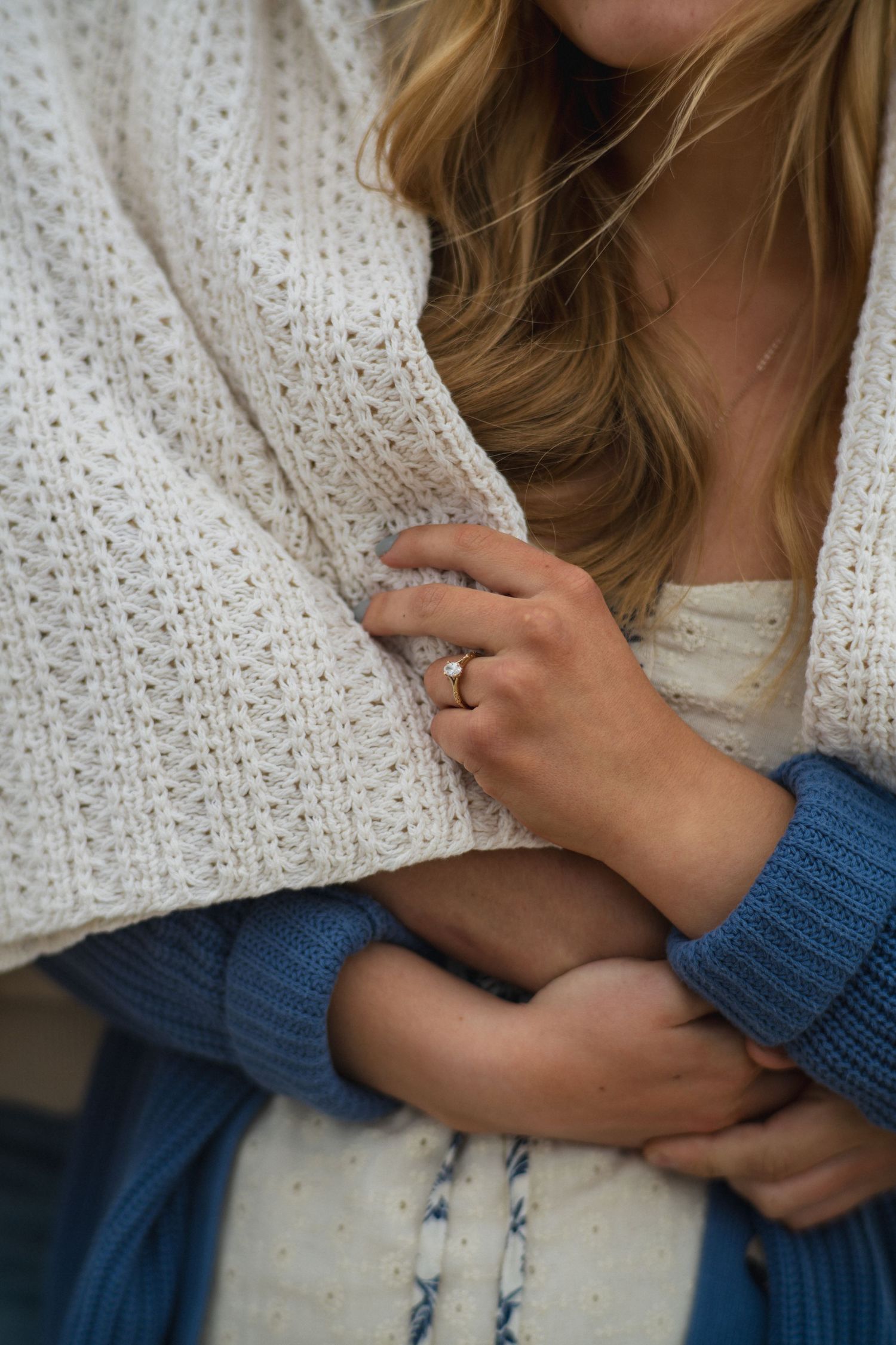 Close-up of a woman in a cream knit cardigan and blue sweater, wearing a diamond ring, hugging herself warmly.