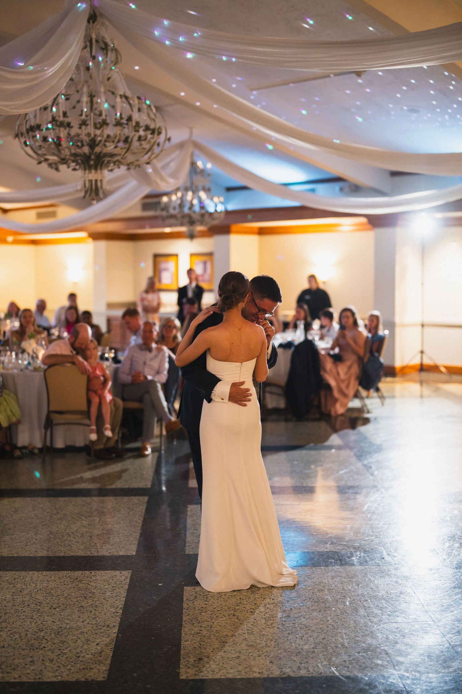 Bride and groom share intimate first dance in elegant ballroom with draped ceiling and crystal chandelier.