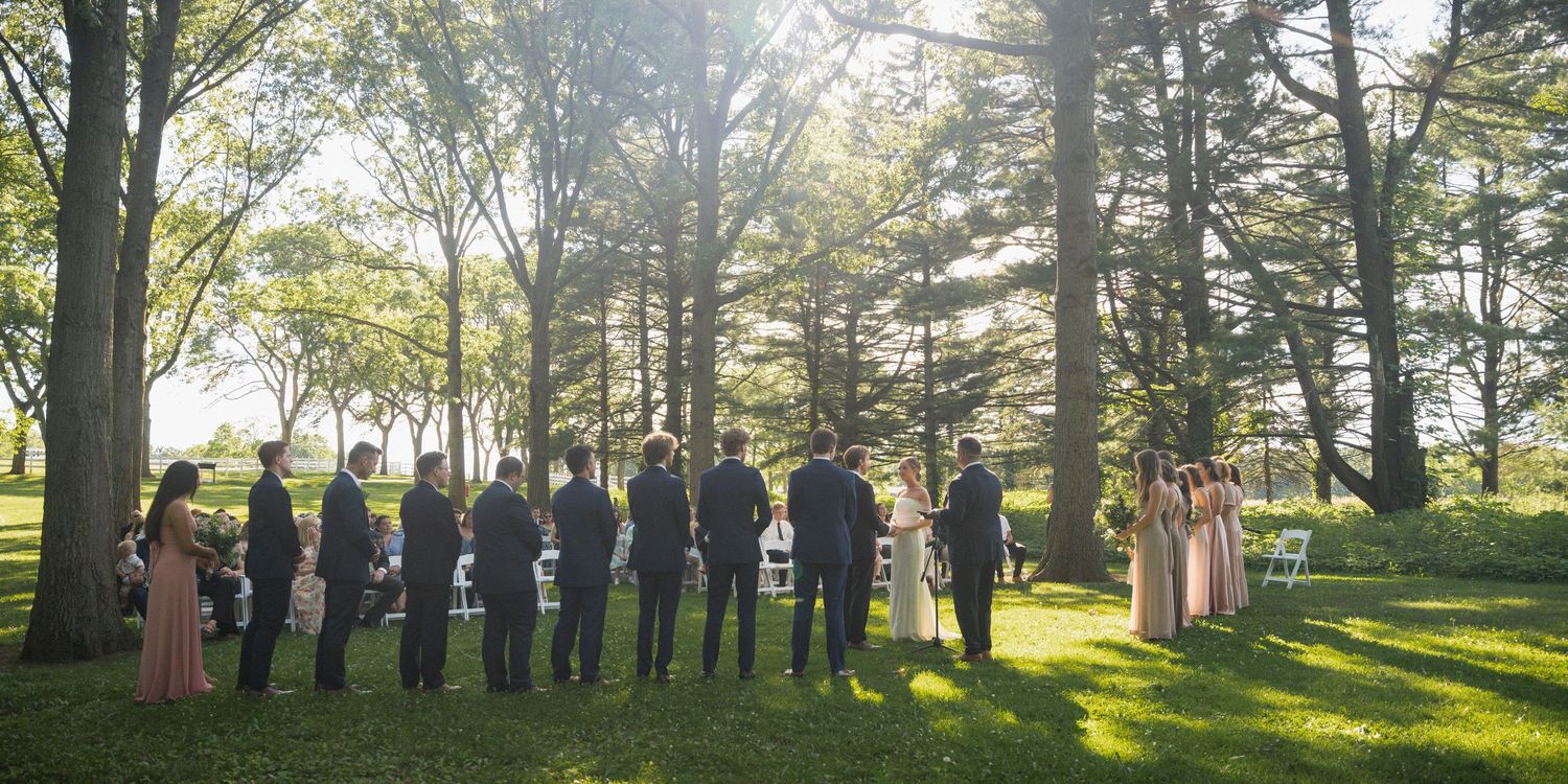 Wide shot of wedding party standing at outdoor ceremony altar surrounded by tall trees.