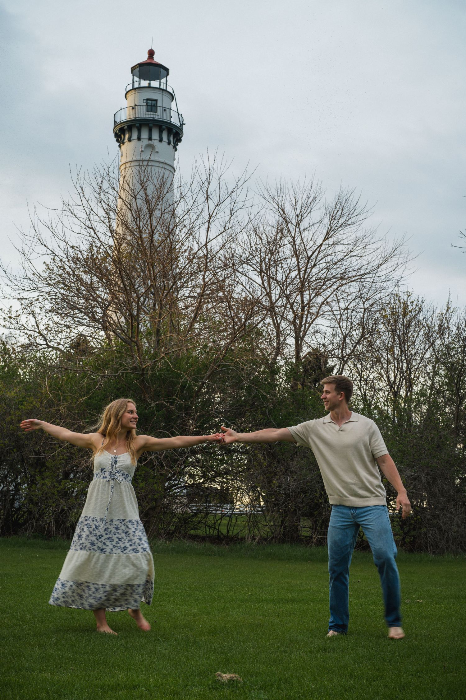 A joyful couple holds hands in a green field with a tall lighthouse and bare trees behind them.