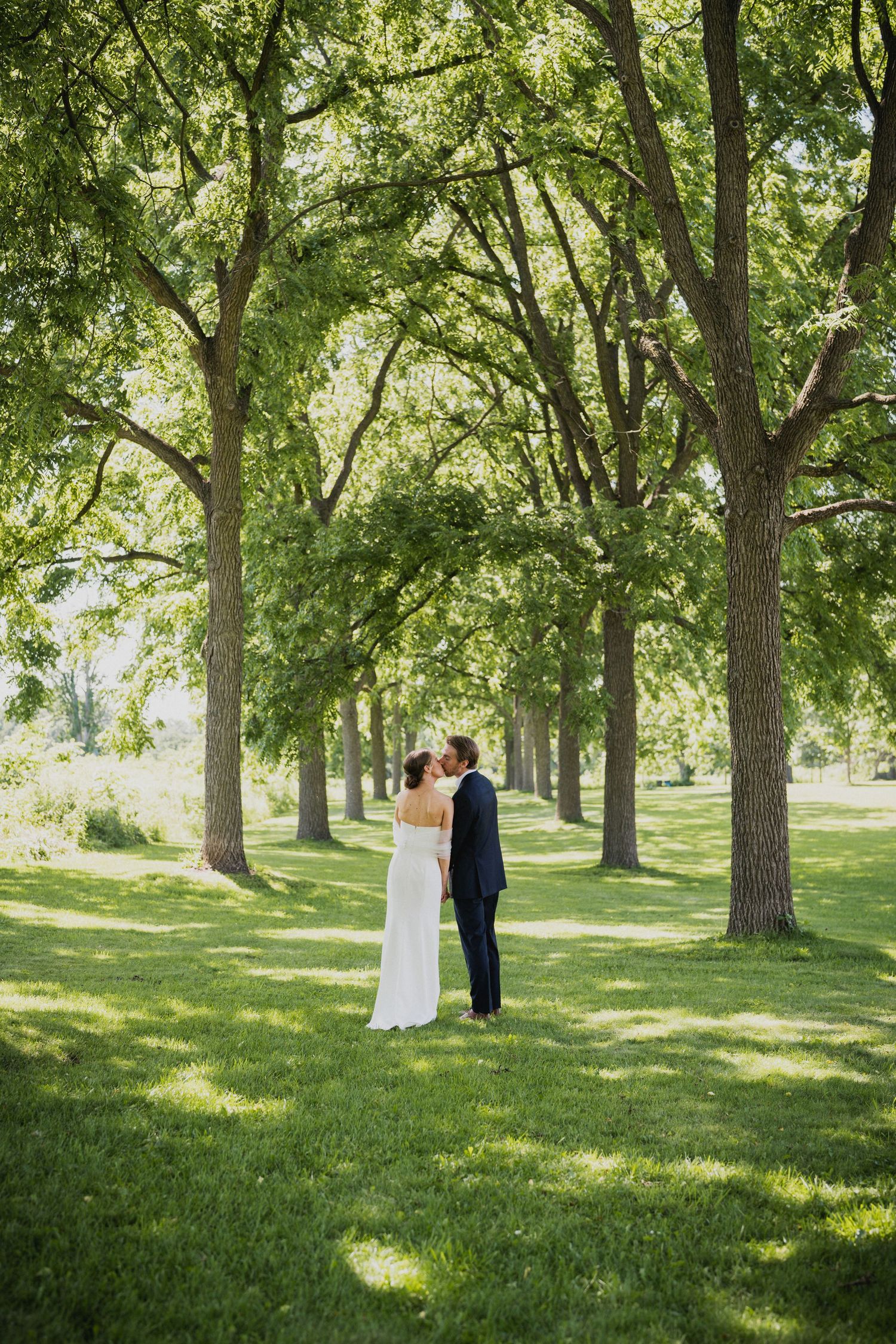 Bride and groom sharing a romantic kiss among a tree-lined lawn on their wedding day.