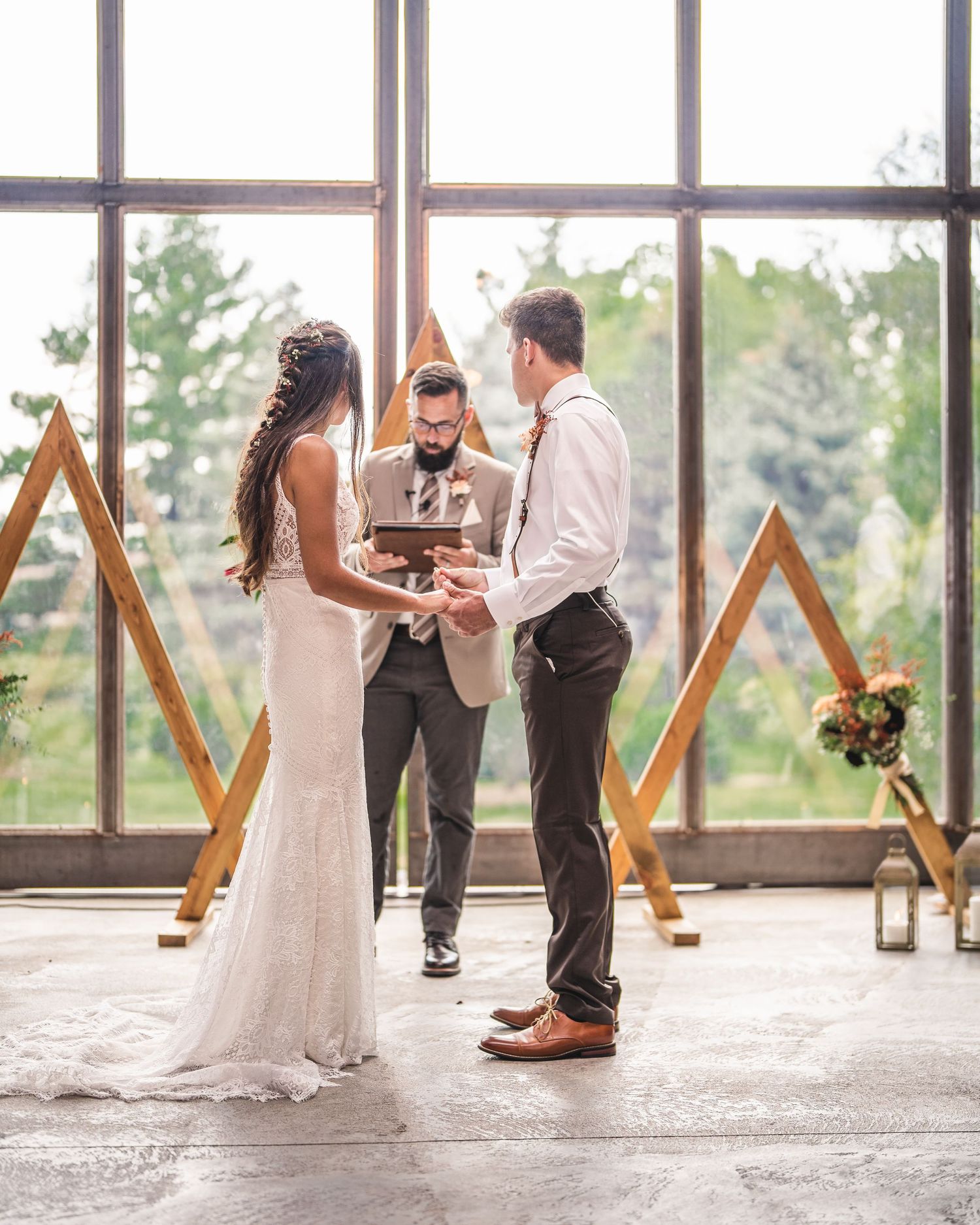 Wedding ceremony in front of geometric wooden arch with lanterns and natural light from floor-to-ceiling windows.