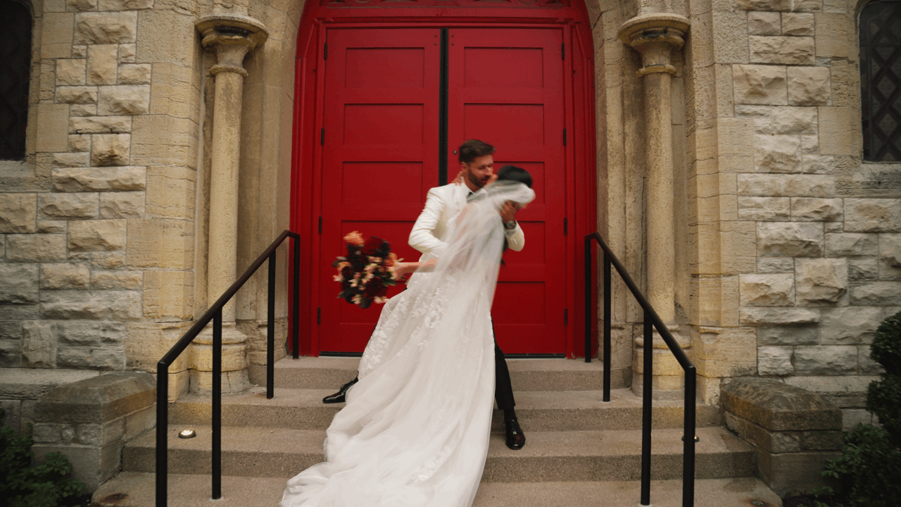 Wedding film of newlyweds dip kissing in front of beautiful red church doors. Then cuts to a romantically lit reception dance scene.