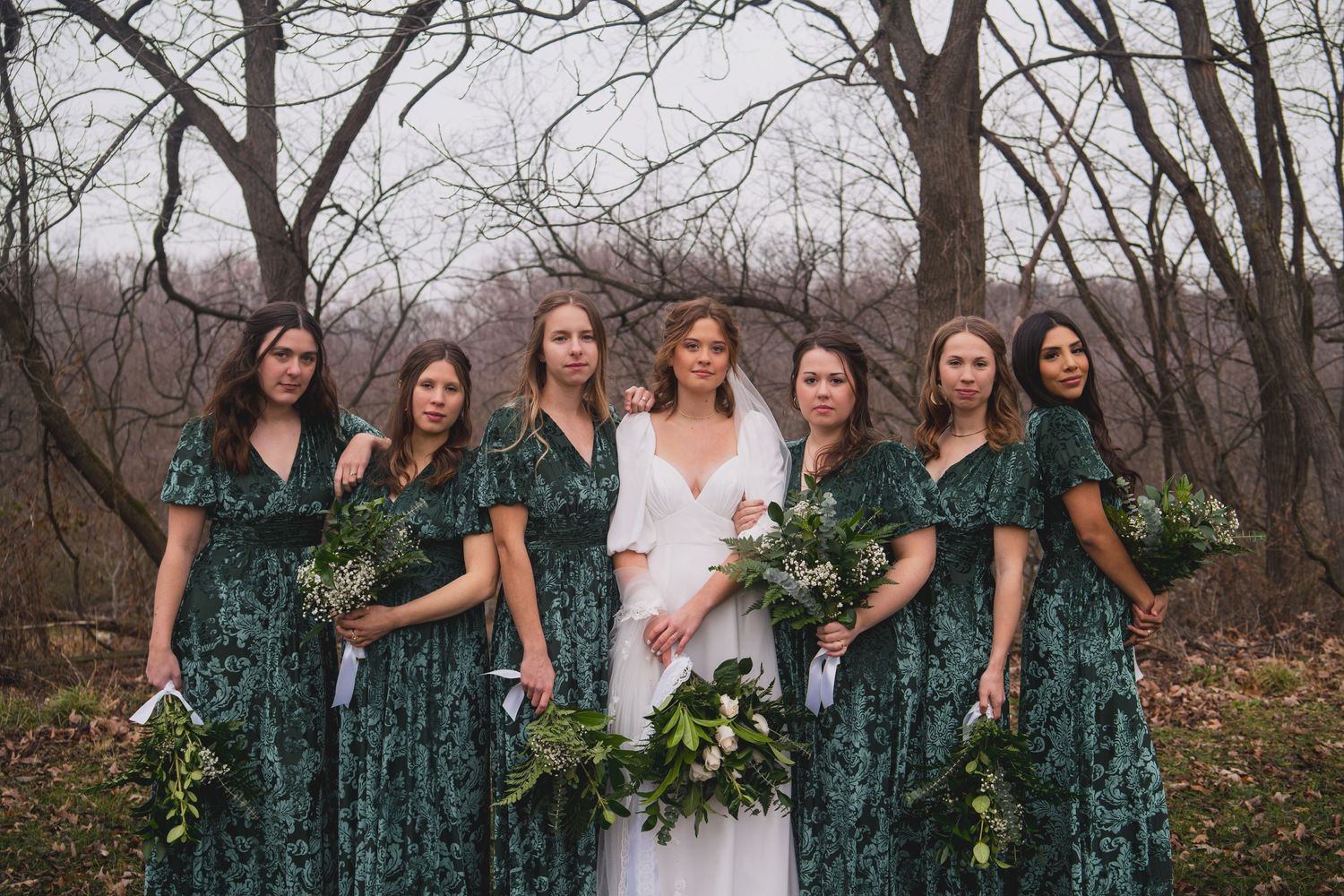 Wedding party in emerald dresses poses with bride holding elegant white and green bouquets.