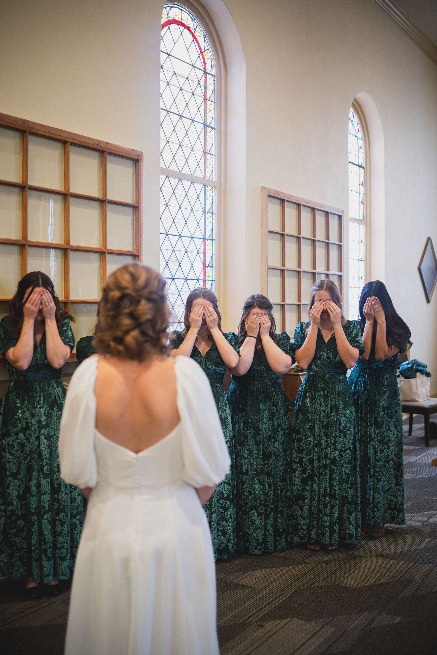 Bridesmaids in dark green dresses hide their faces while bride in white stands at the end.