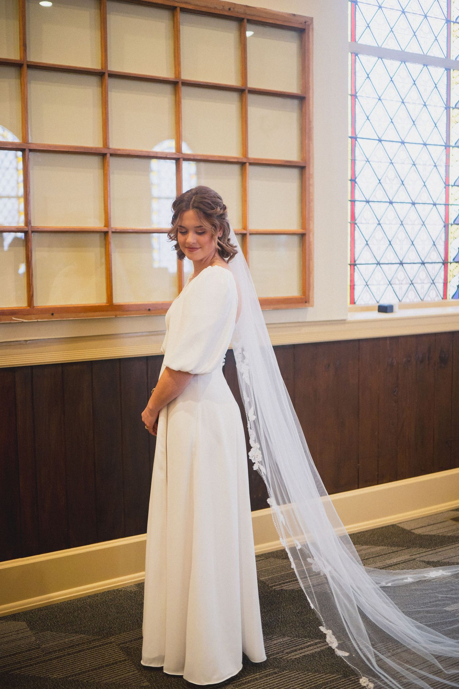 Elegant bride in white dress with flowing veil poses by church window.