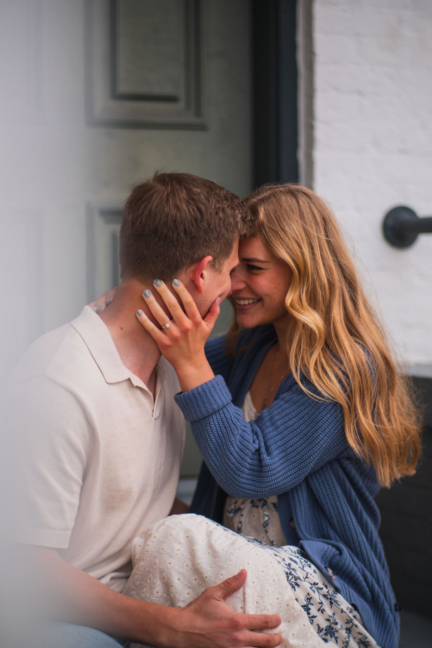 Smiling woman tenderly holds man's face as they share an intimate moment on a porch.