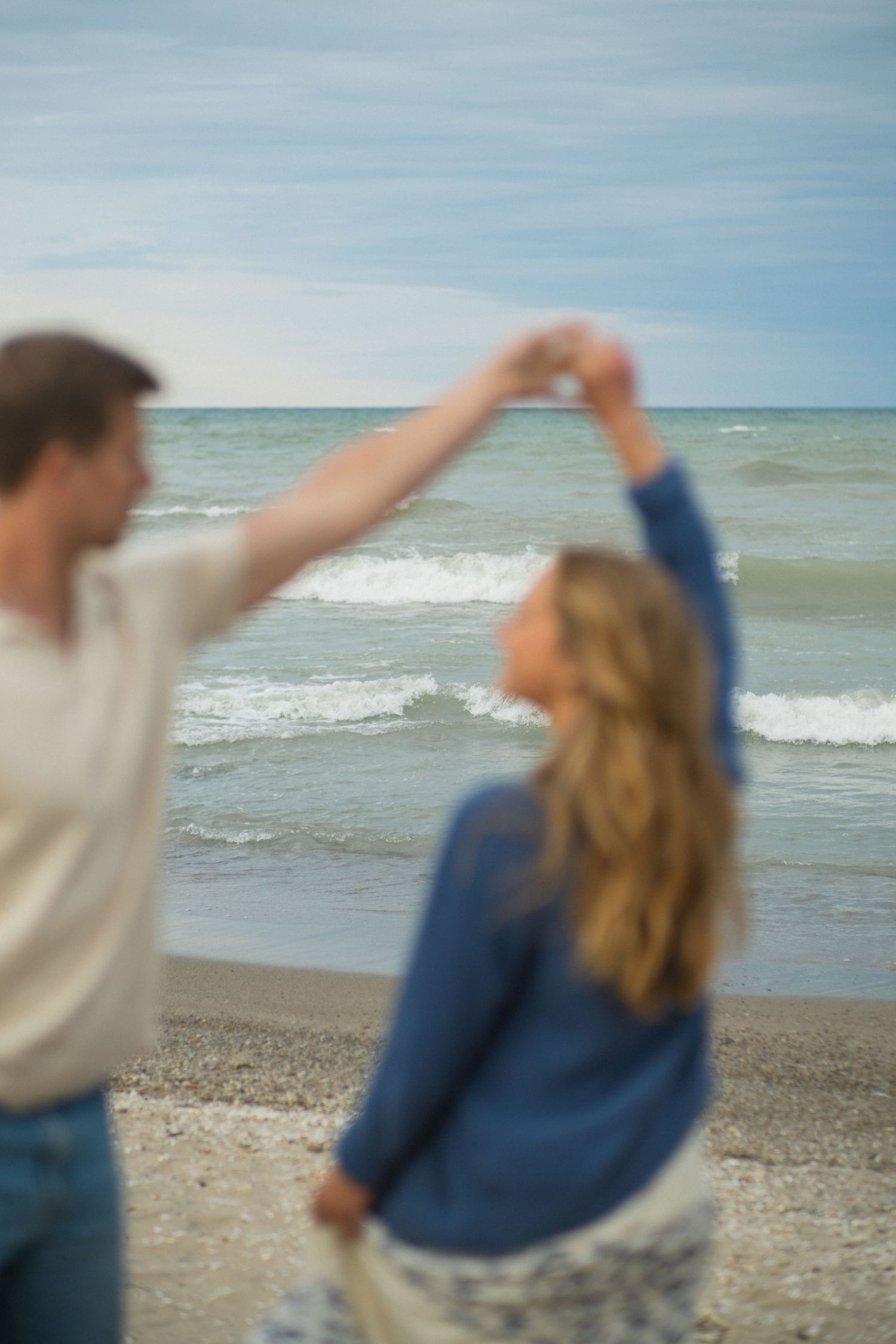 Couple dancing romantically on a beach shoreline with ocean waves in the background on a cloudy day.