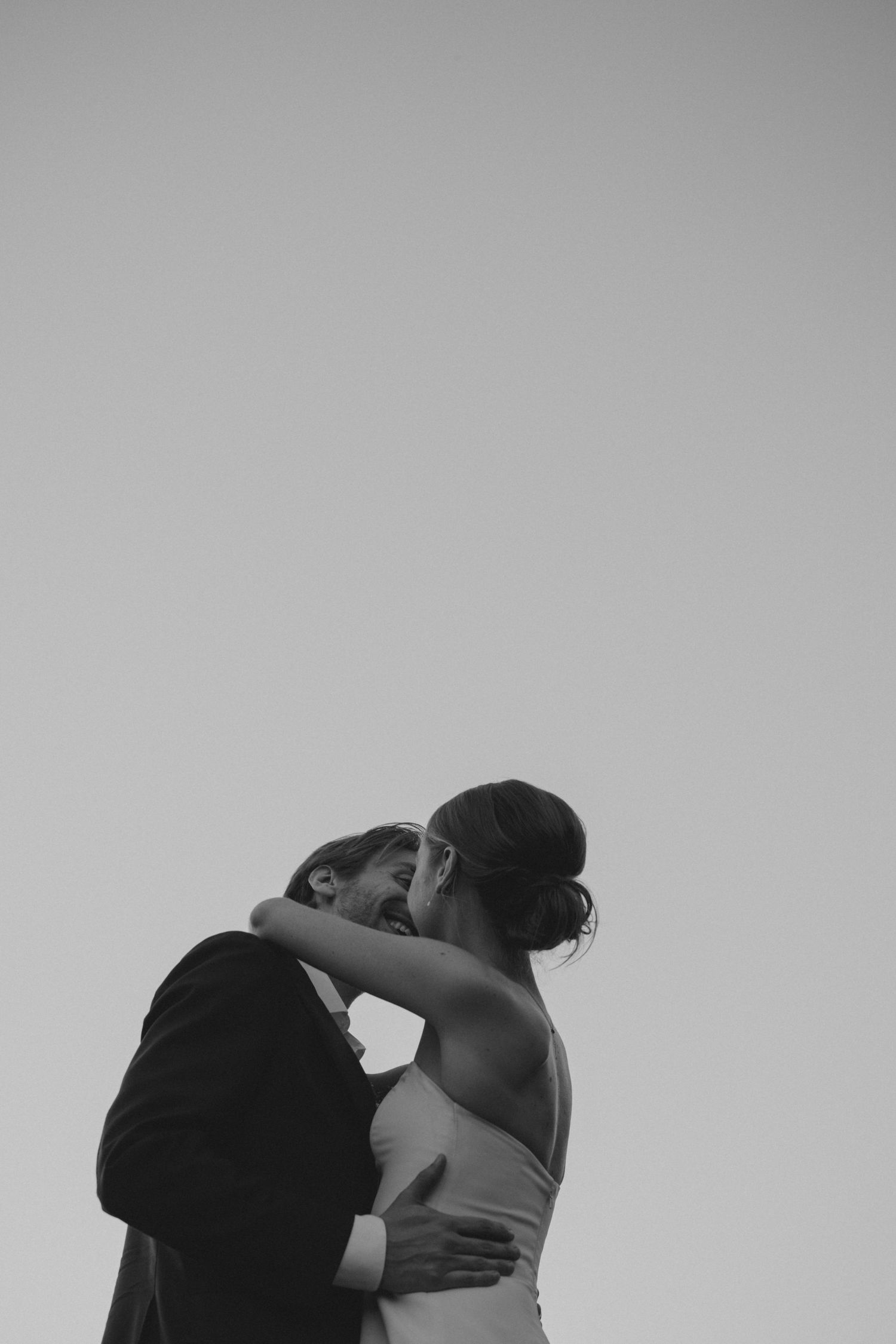 Black and white photo of a couple sharing a romantic kiss, the bride in a sleeveless gown embracing the groom in a suit.