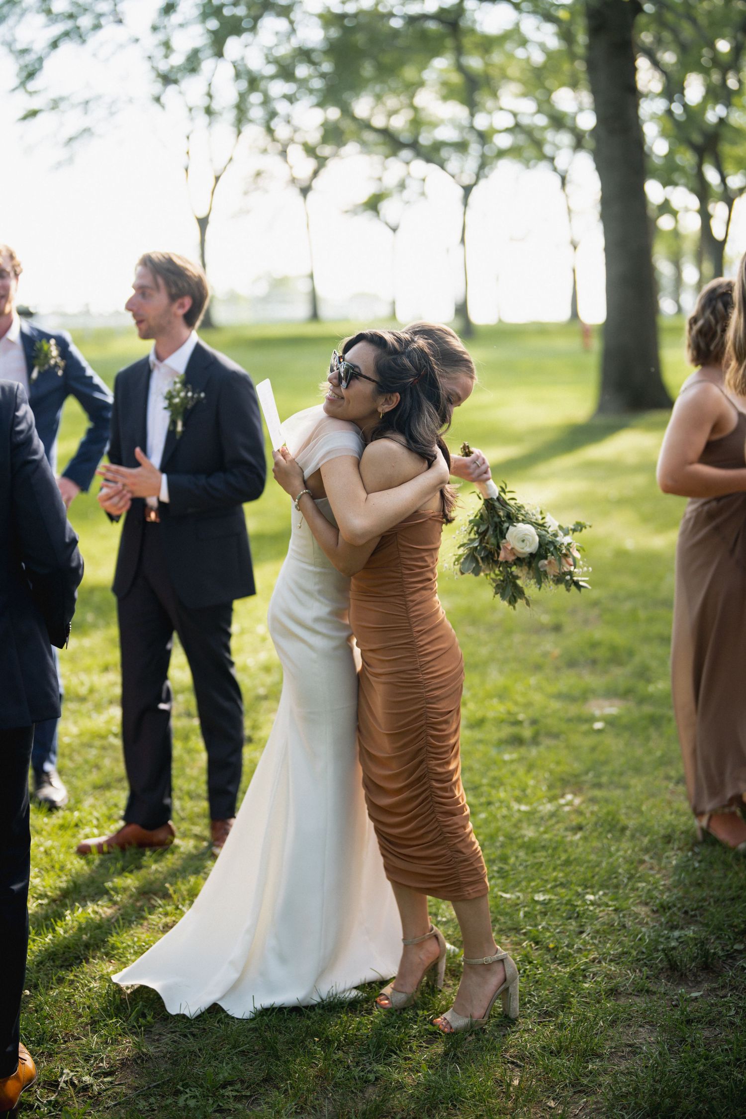 Bride hugging a guest in a brown ruched dress outdoors at a wedding reception on a sunny day.