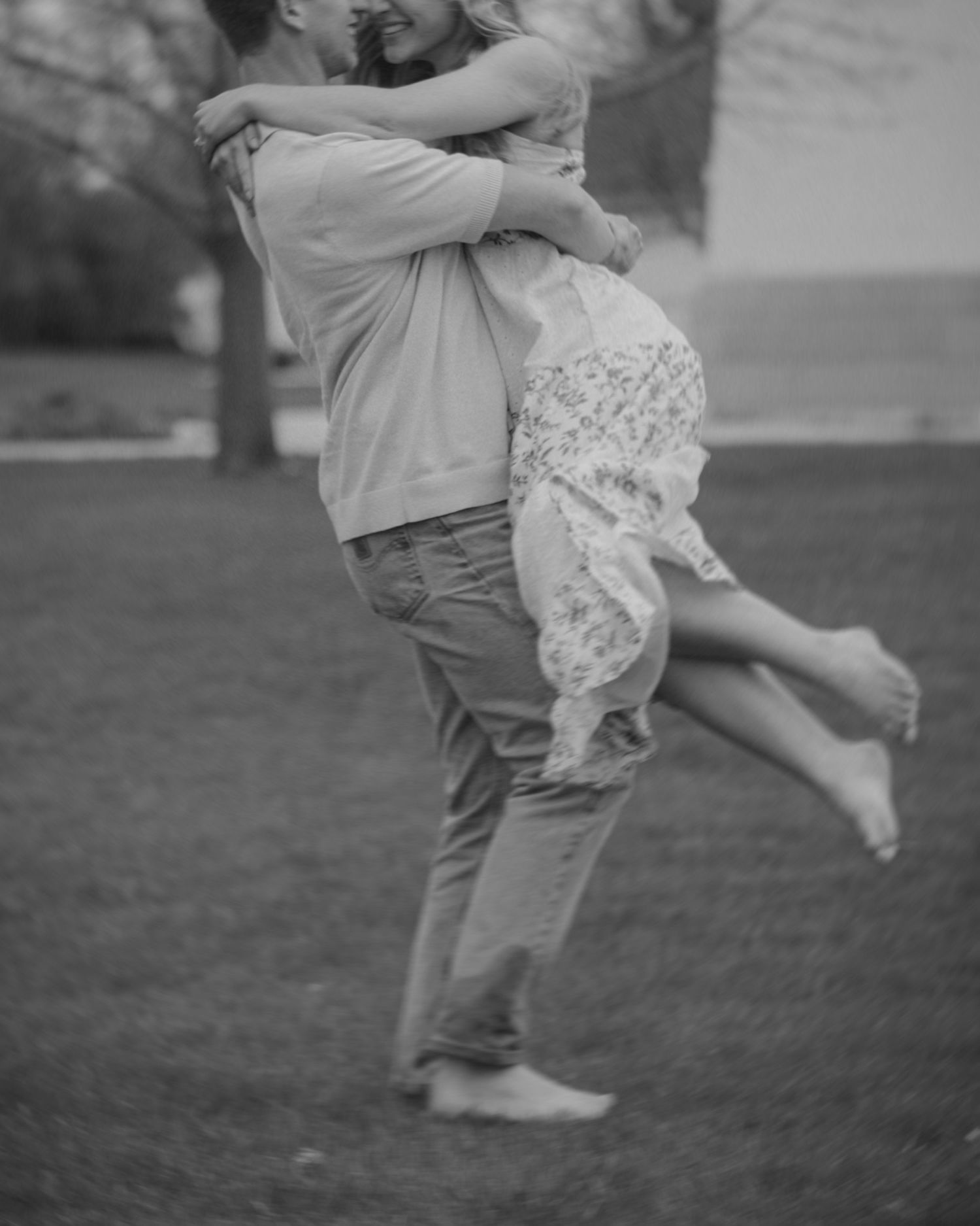 Black and white photo of a couple embracing outdoors, man lifting woman in floral dress while she wraps arms around him.
