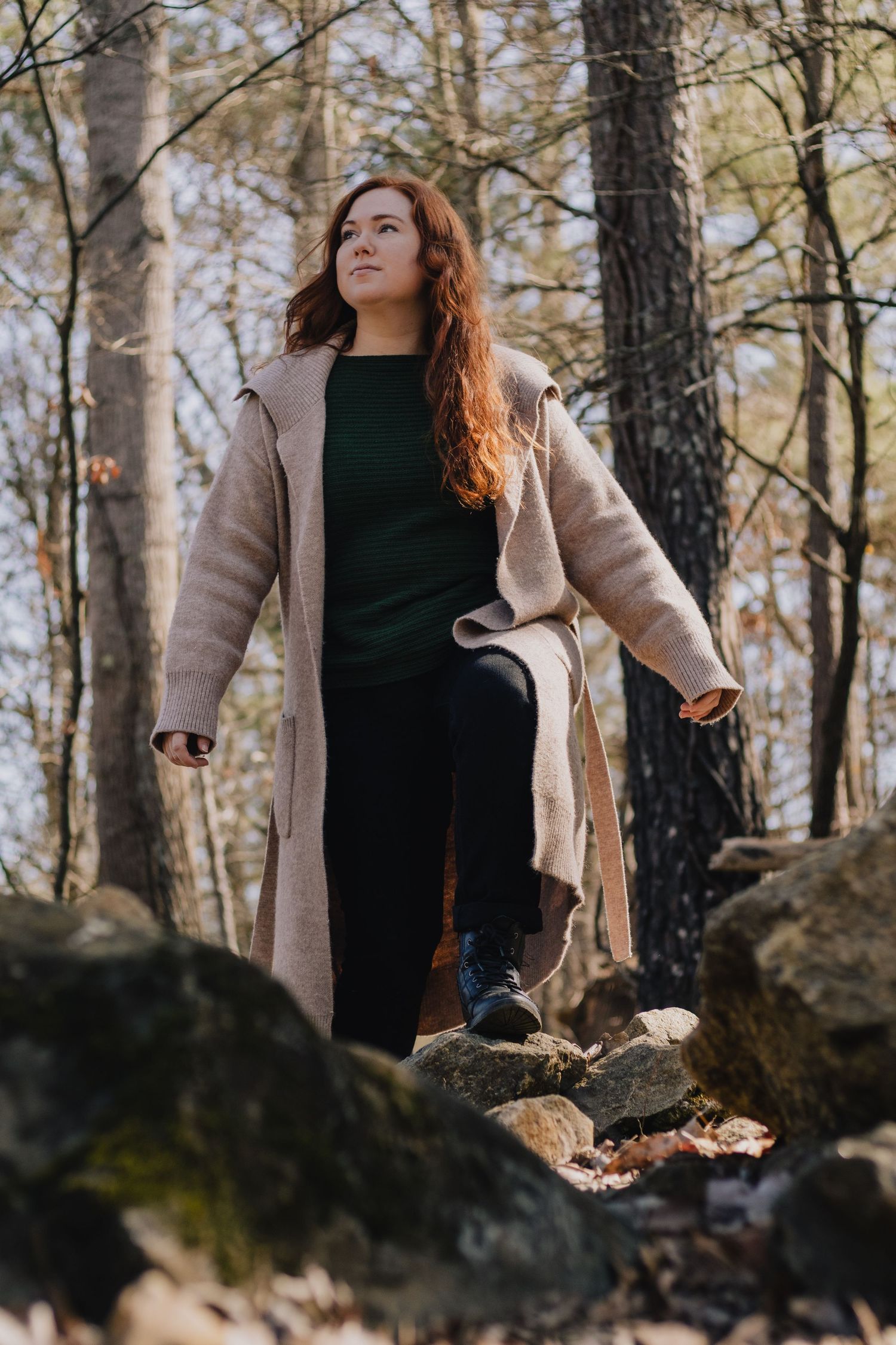 Figure in beige coat and dark clothing stands on rocky forest terrain looking into distance.