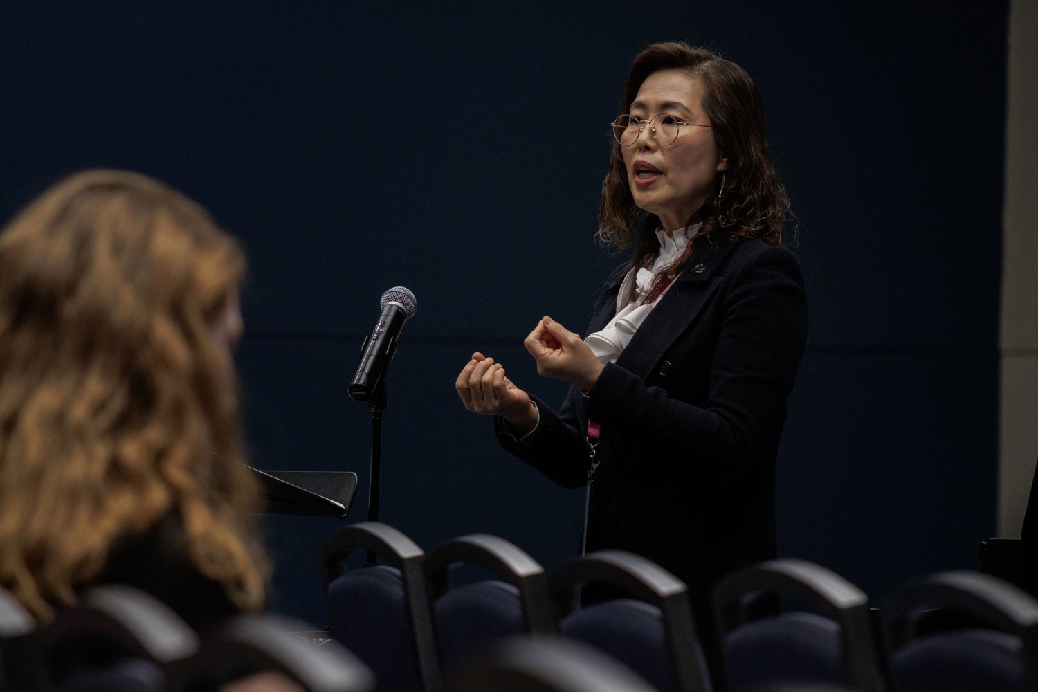 A speaker gestures while giving a presentation in a dark auditorium setting.