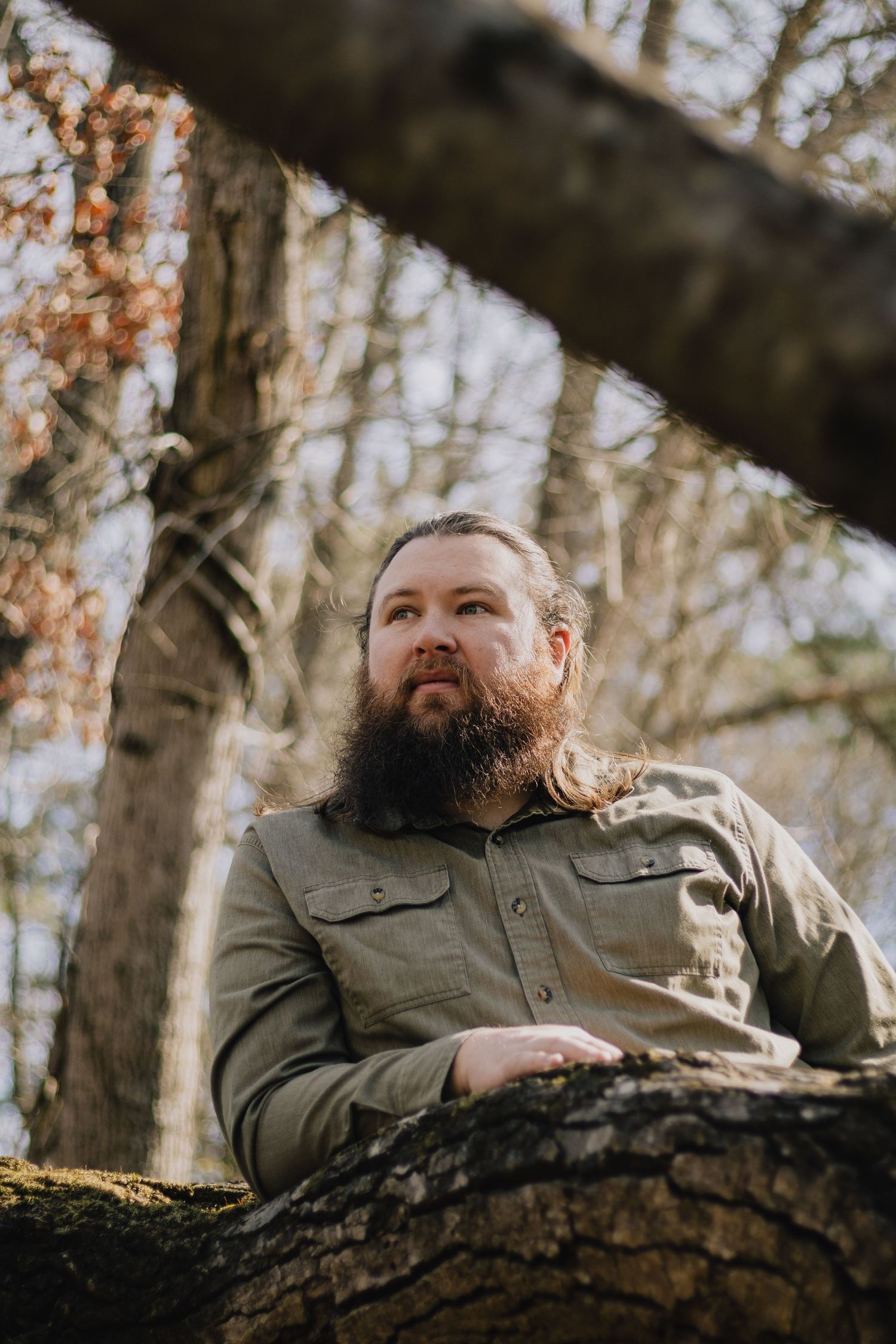 Person with beard rests against tree trunk in autumn forest setting.