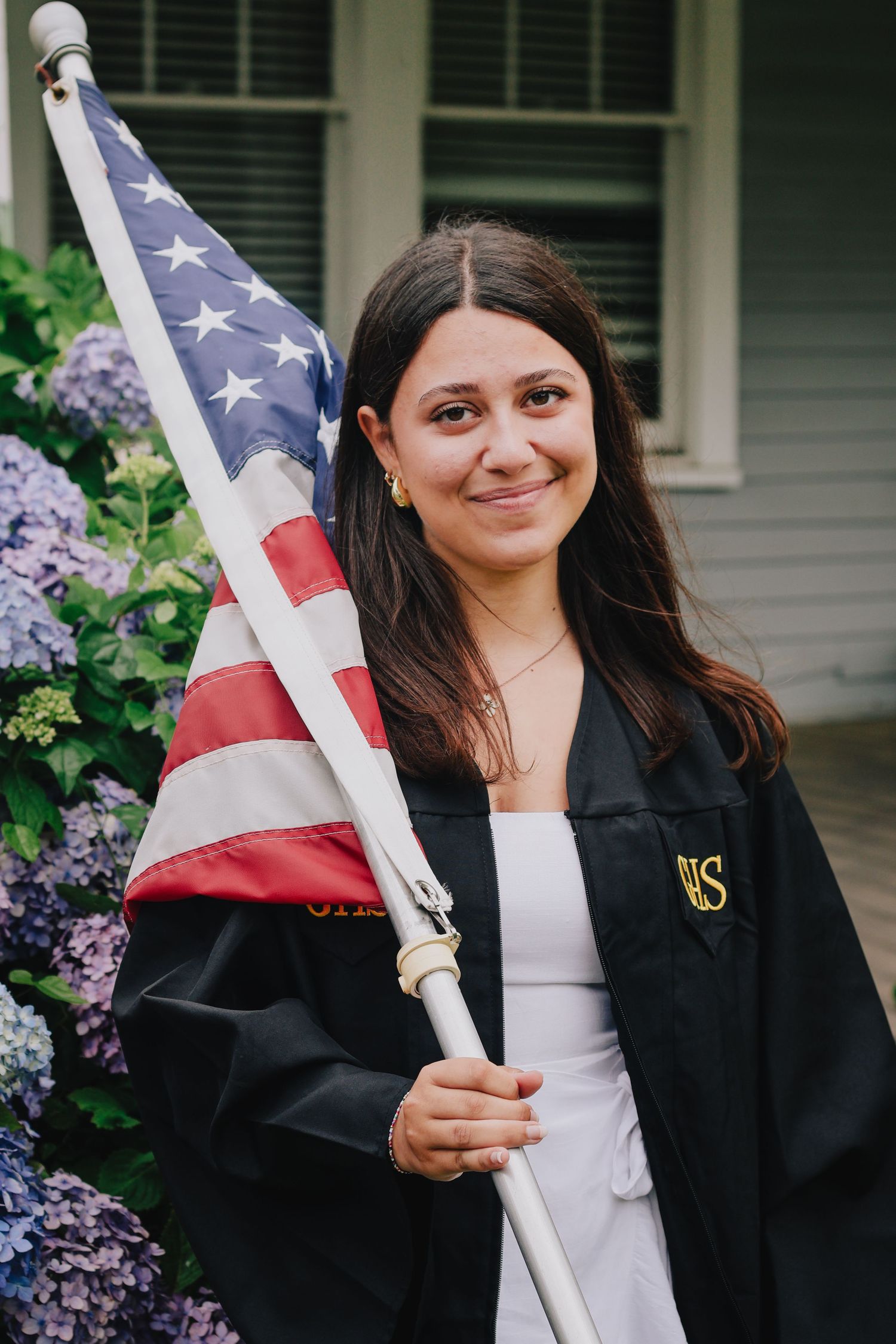 A graduate in academic regalia holding an American flag near blooming lilac bushes.