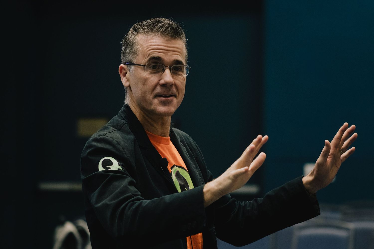 A speaker in casual attire makes an expansive gesture with both hands during a presentation.