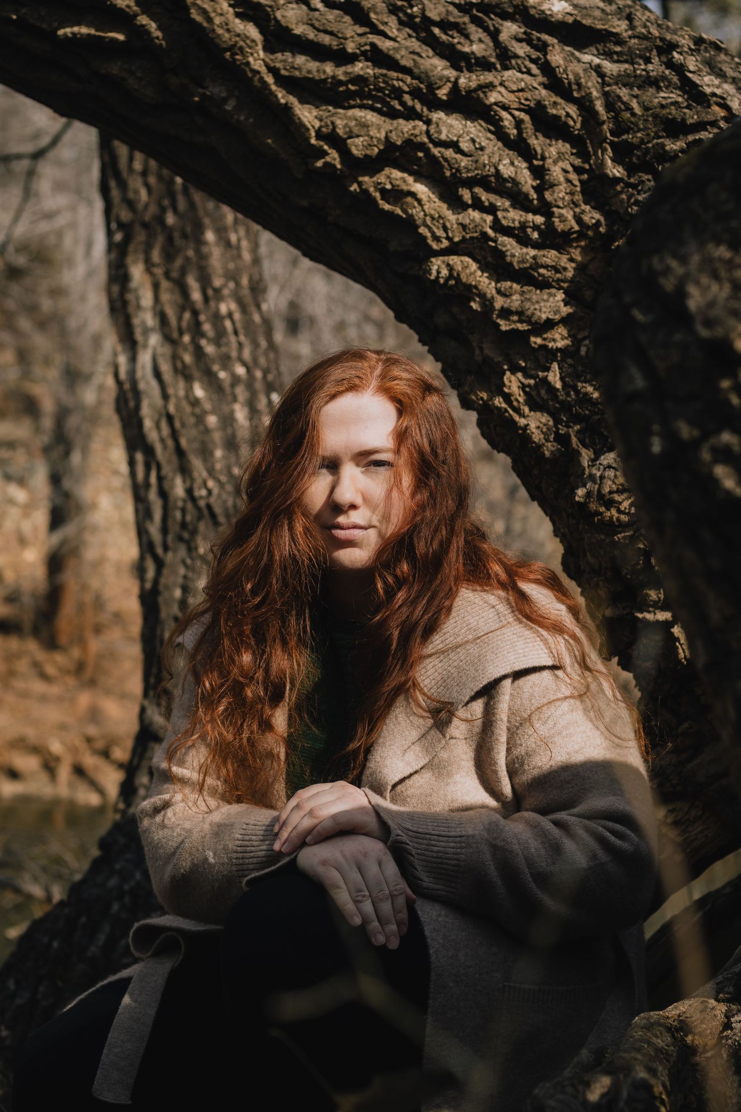 Person with long red hair sits under a large tree trunk wearing a brown coat in a moody forest setting.